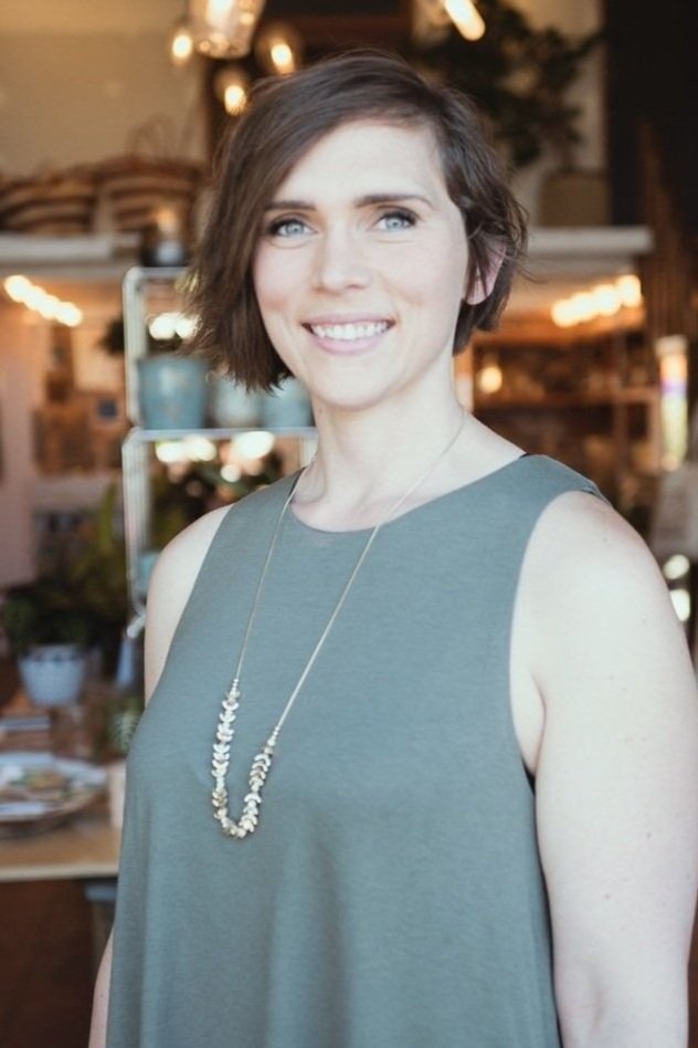 A woman with short brown hair and blue eyes smiling at the camera inside a warmly lit cafe or restaurant, wearing a sleeveless gray top and a long necklace.