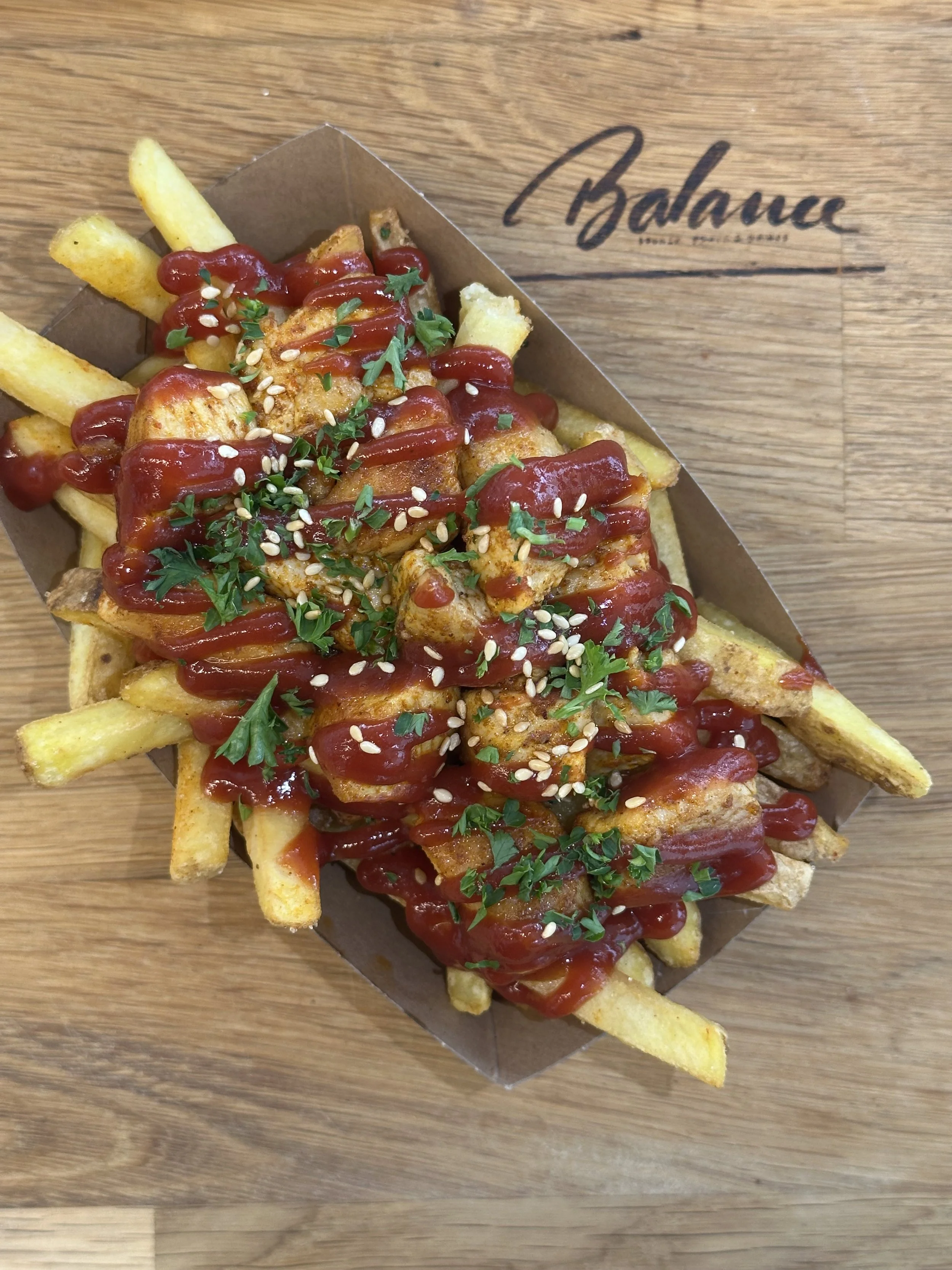 Pommes Frites mit Hähnchenstücken, Ketchup, Petersilie und Sesamsamen in Pappschale auf Holzplatte.