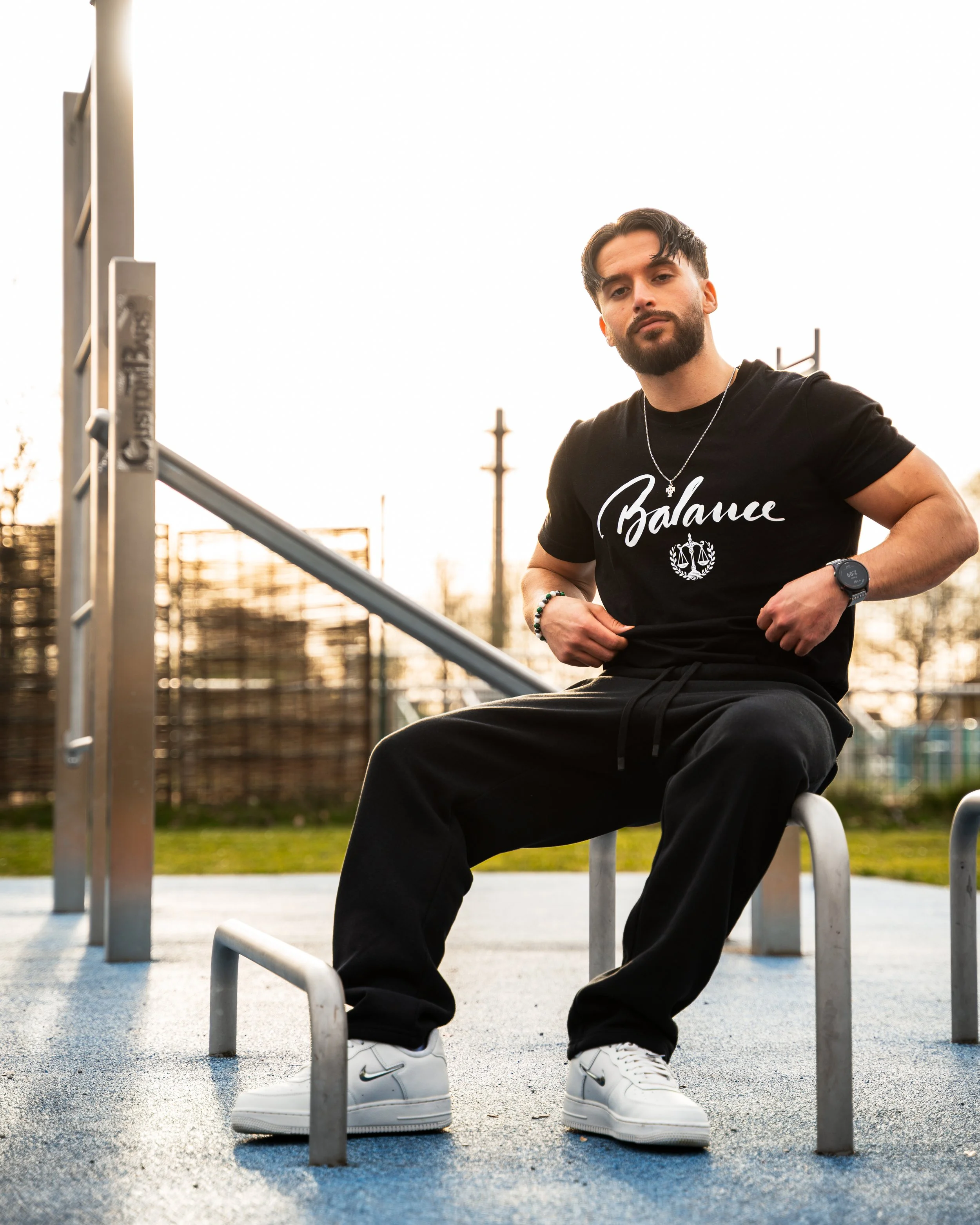 Mann sitzt auf einer Bank im Freien bei Sonnenuntergang, trägt ein schwarzes T-Shirt mit dem Wort 'Balance' und einer Waage, weiße Nike-Sneaker, eine Smartwatch und Schmuck, im Hintergrund eine Metallstruktur und Bäume.