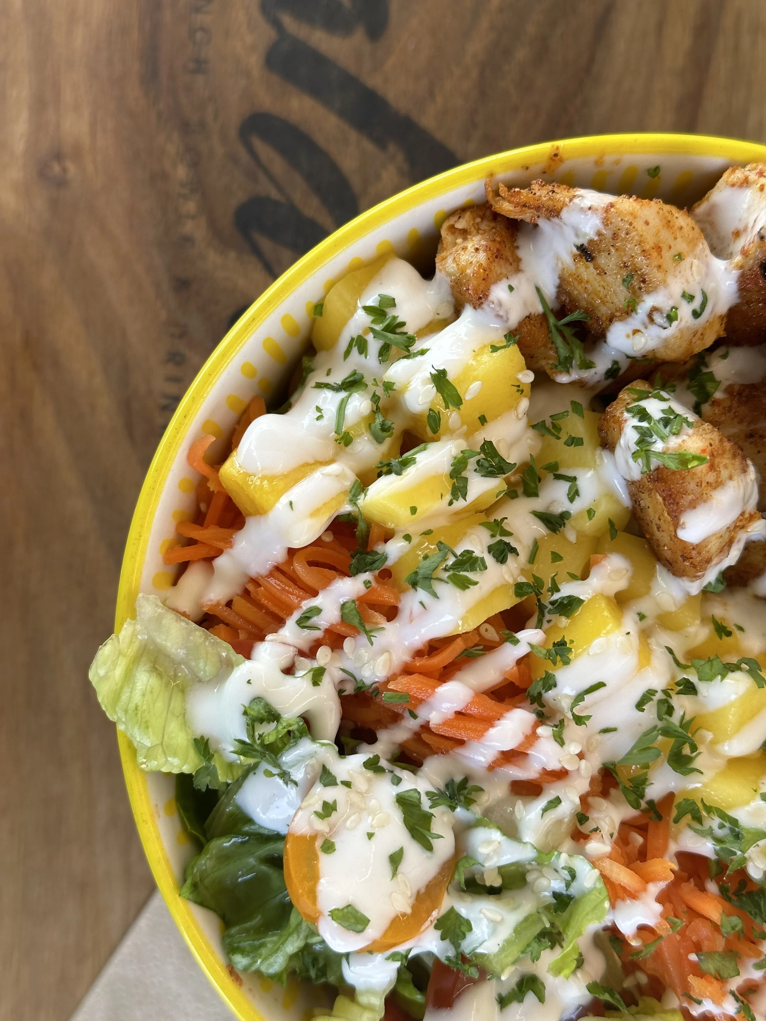 Bunter Salat mit Tomaten, Karotten, Gurken, Paprika, Mango, knusprigem Hühnchen, Joghurtdressing und frischer Petersilie in einer gelben Schale auf einem Holztisch.