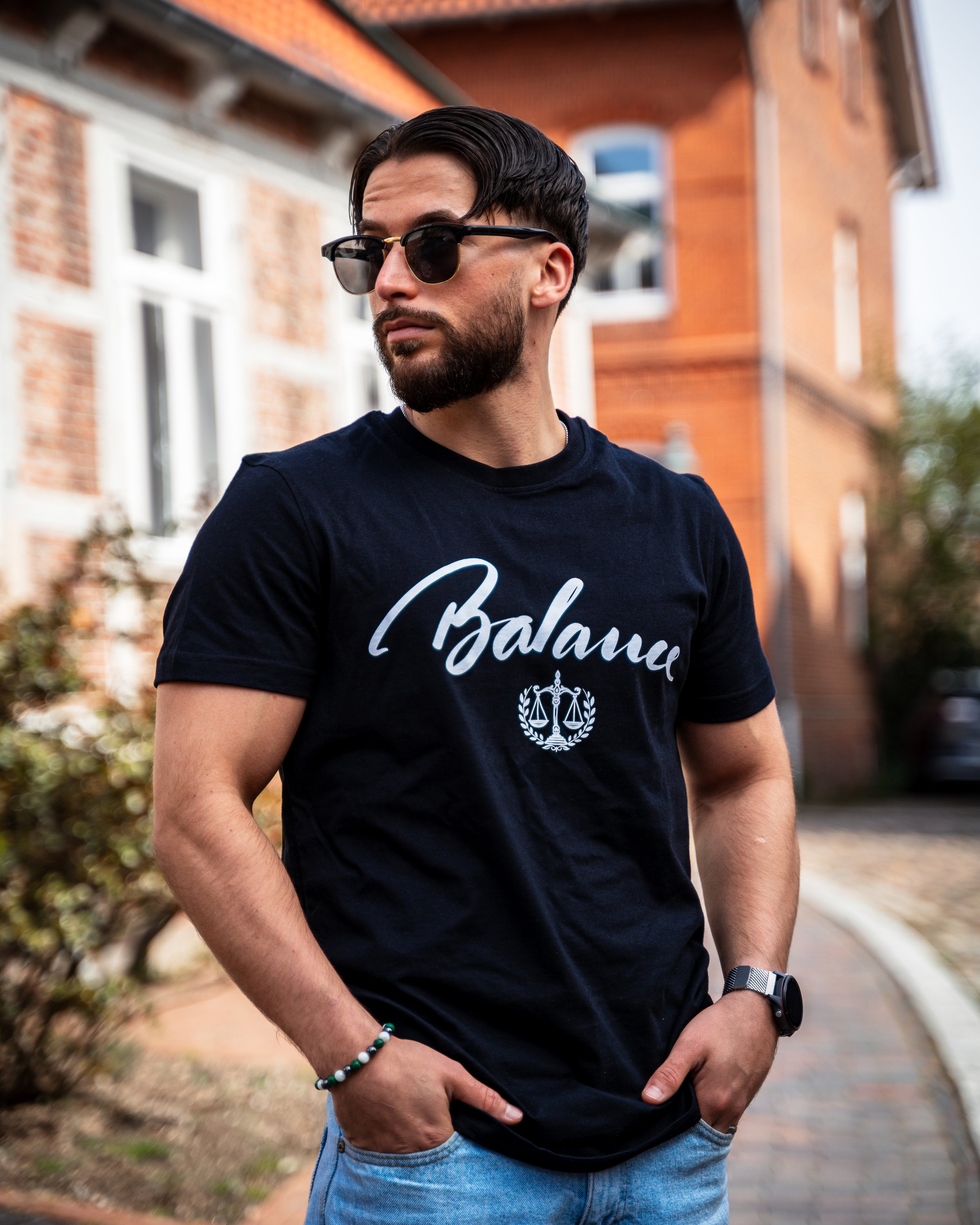Ein junger Mann mit dunklen Haaren, Bart, Sonnenbrille und schwarzem T-Shirt steht vor einem Backstein-Gebäude. Er trägt eine Armbanduhr und ein Armband und hat die Hände in den Taschen. Im Hintergrund sind Bäume und eine Straße zu sehen.