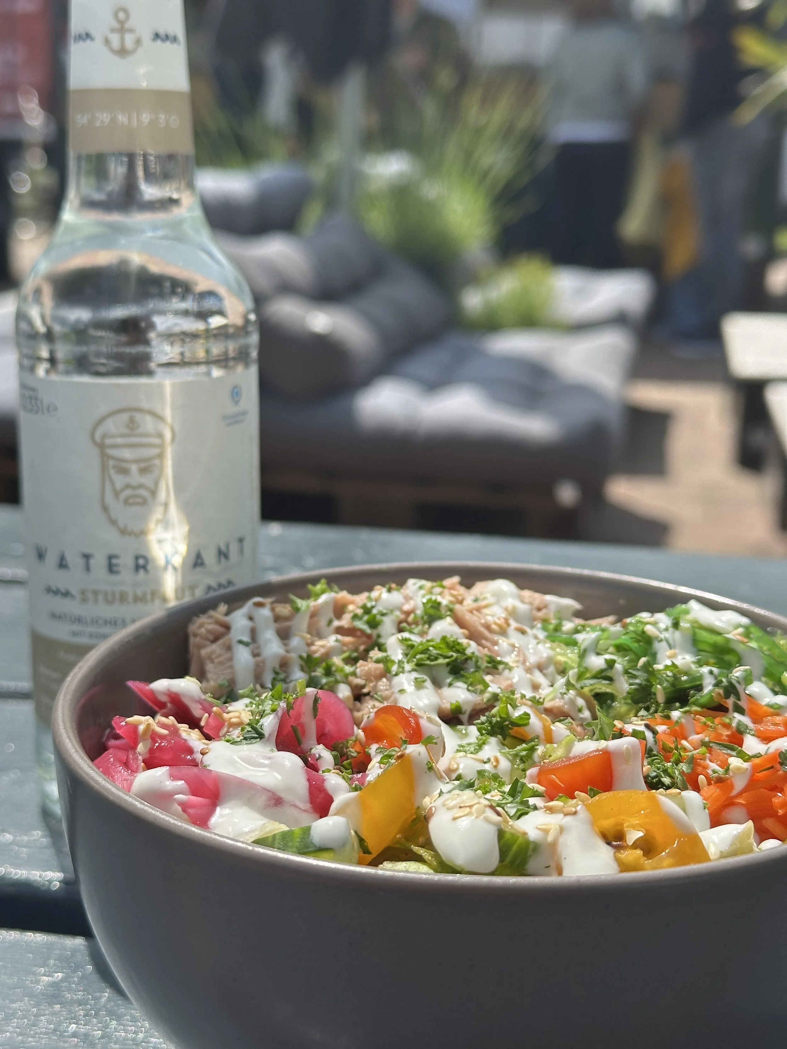 Eine Schüssel mit buntem Salat, garniert mit Sesamsamen und einem Dressing, auf einem Tisch im Freien, im Hintergrund eine Wasserflasche und eine Gartenlounge.