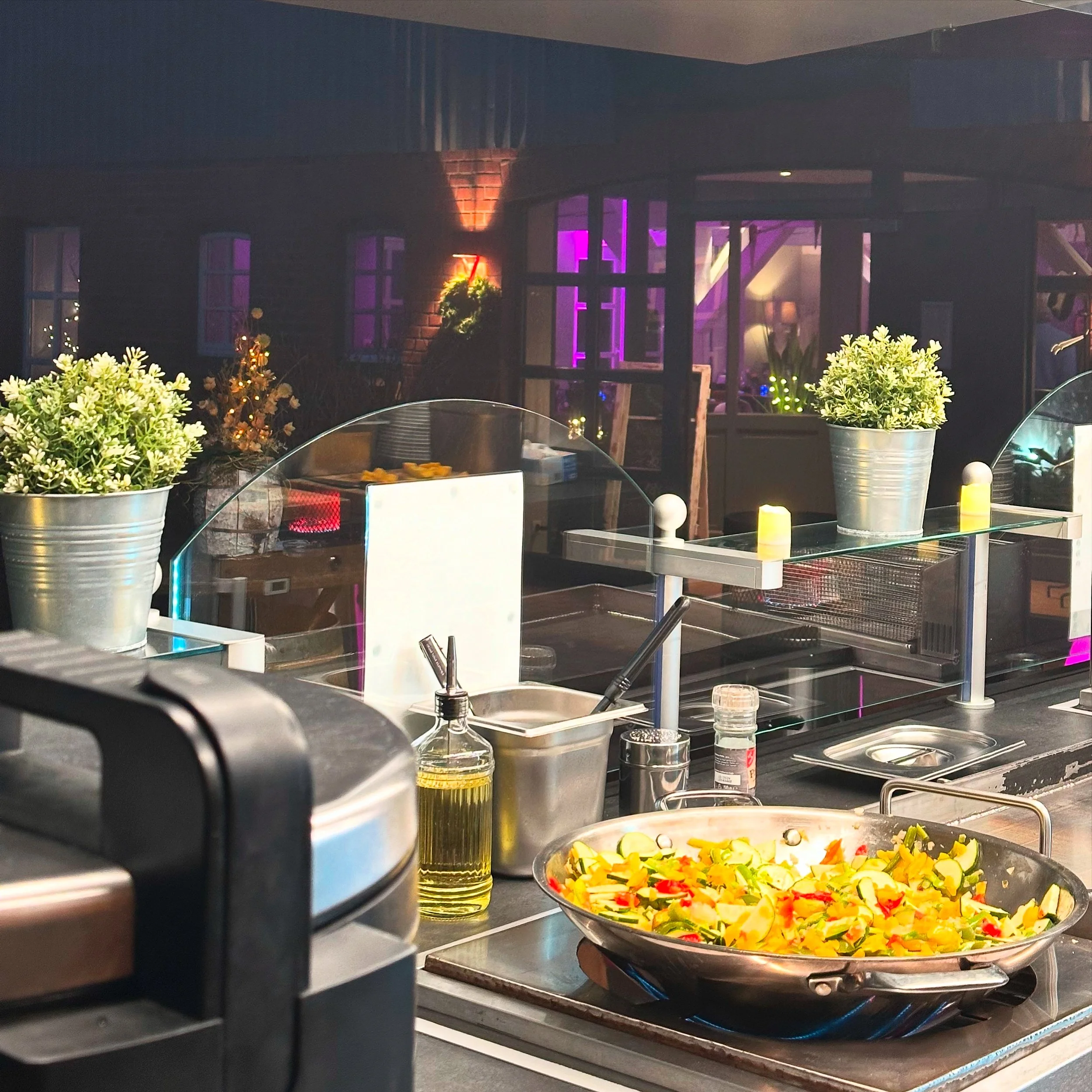 Blick auf eine Restaurantküche bei Nacht mit einem Wok voller leckerer Gemüsescheiben, Pflanzen in Töpfen, Küchenutensilien und einer Maschine im Vordergrund, sowie einem Fenster mit Blick auf ein beleuchtetes restaurantartiges Gebäude im Hintergrund.