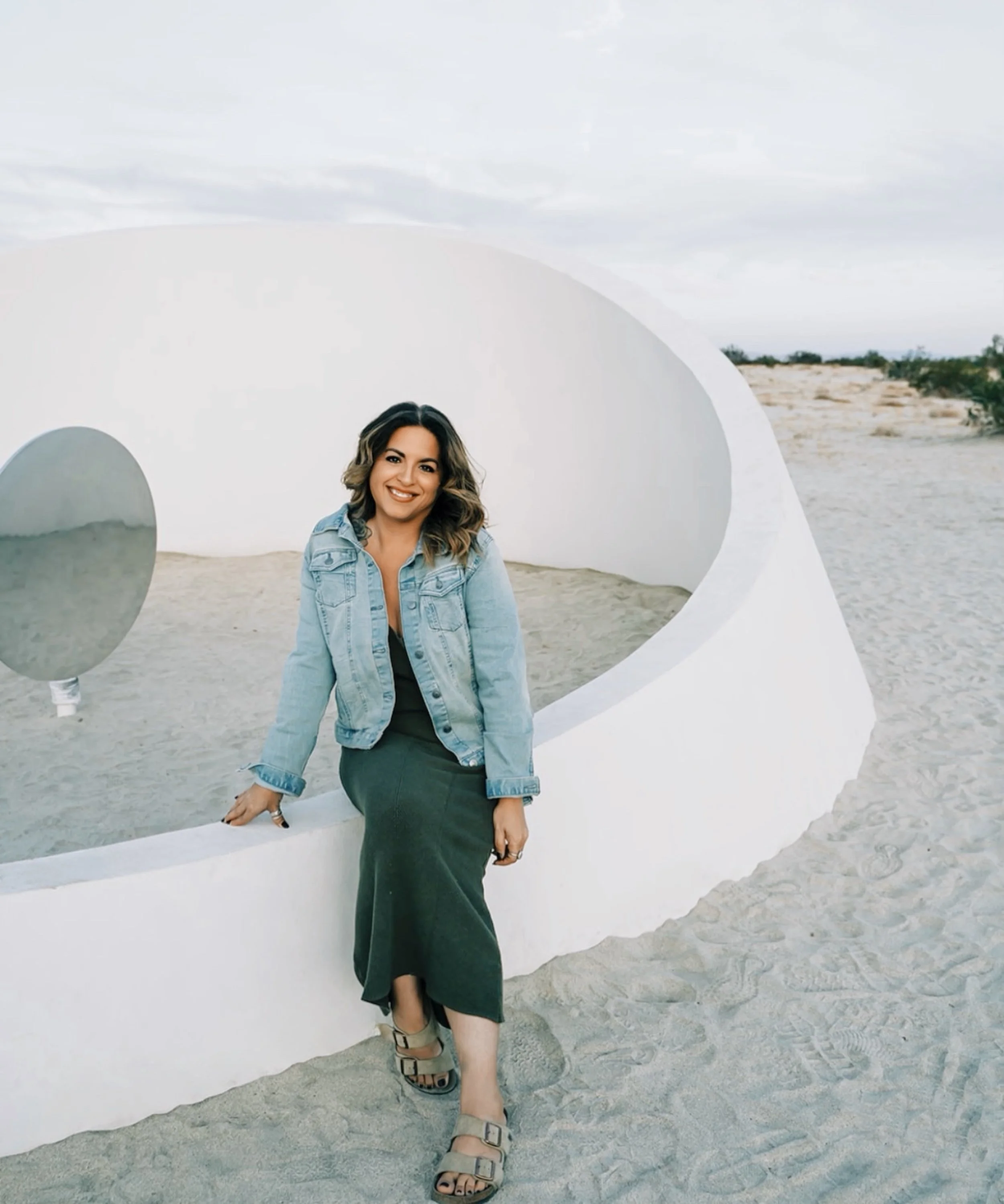 Woman sitting on white curved structure in a desert with a mirror nearby and cloudy sky in the background.