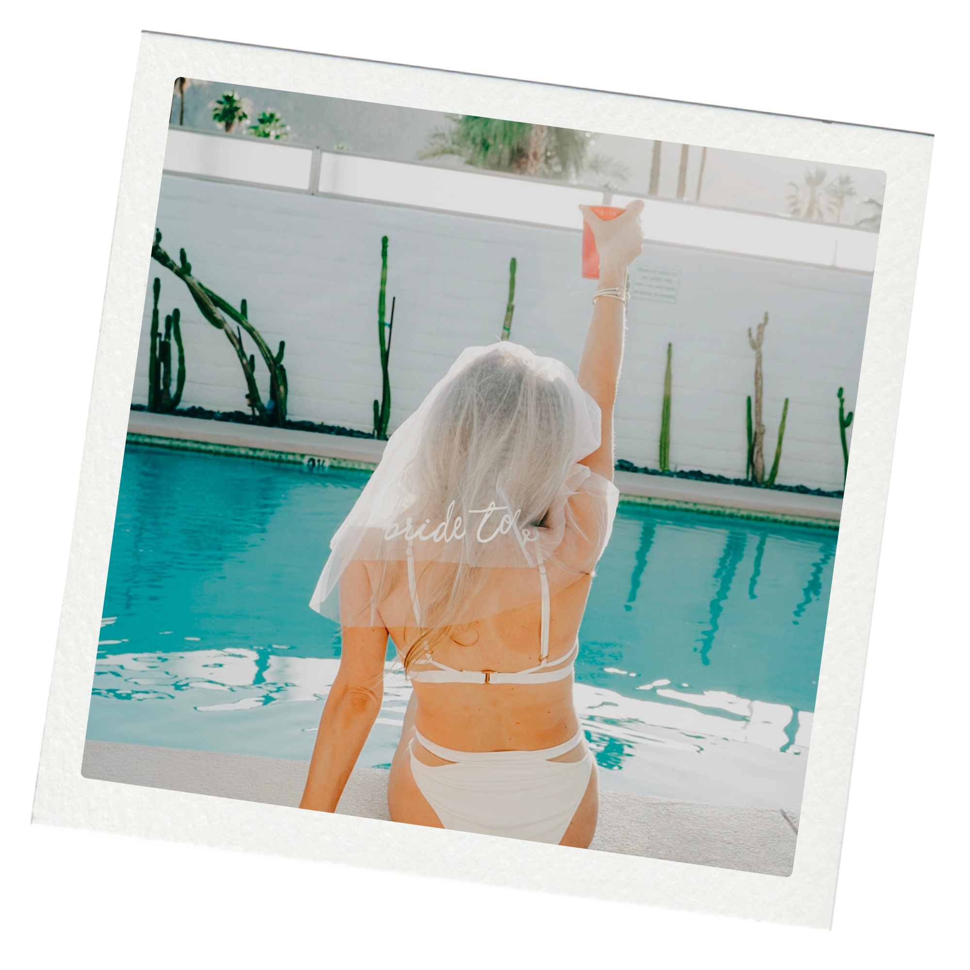 A woman by a pool wearing a bridal veil with the words 'bride to be' on it, taking a selfie with her smartphone.