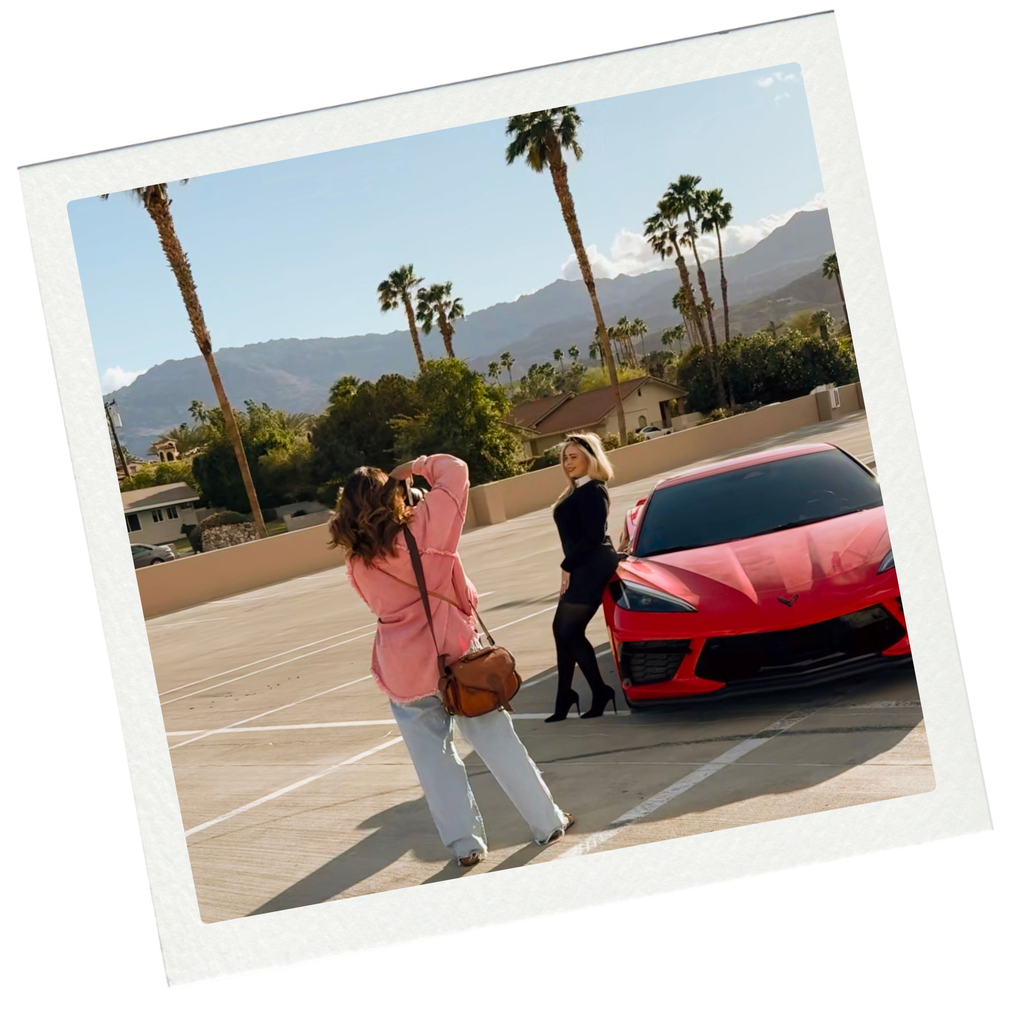 The image shows a woman in a black dress and high heels standing next to a red sports car in an outdoor parking lot with a backdrop of palm trees and mountains during daytime. A second woman, taking a photo, is wearing a pink jacket, white pants, and carrying a brown shoulder bag.
