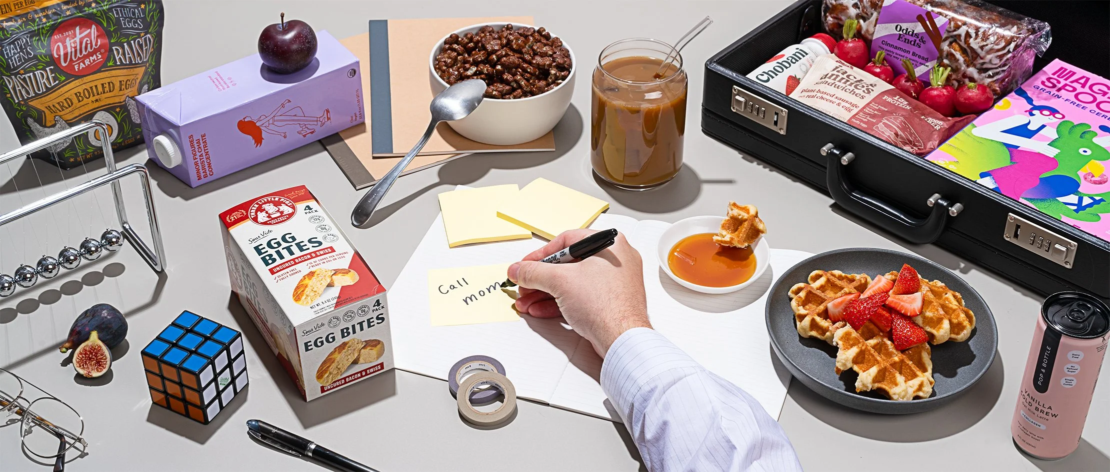 A cluttered desk with breakfast items like waffles topped with strawberries, a bowl of cereal, a plate of waffles with strawberries, and a cup of honey. There are snacks like egg bites, a chocolate bar, cherries, apples, and a bag of raisins. Office supplies include sticky notes, a black pen, scissors, a Rubik's cube, and a roll of tape. A hand is writing "Call mom" on a notepad. There's a drink, a black briefcase open with various snack packages inside, and a pink canned coffee. Also, a glass of iced coffee, a bowl of cooked beef, a small bowl of honey, and a pair of glasses on the desk.