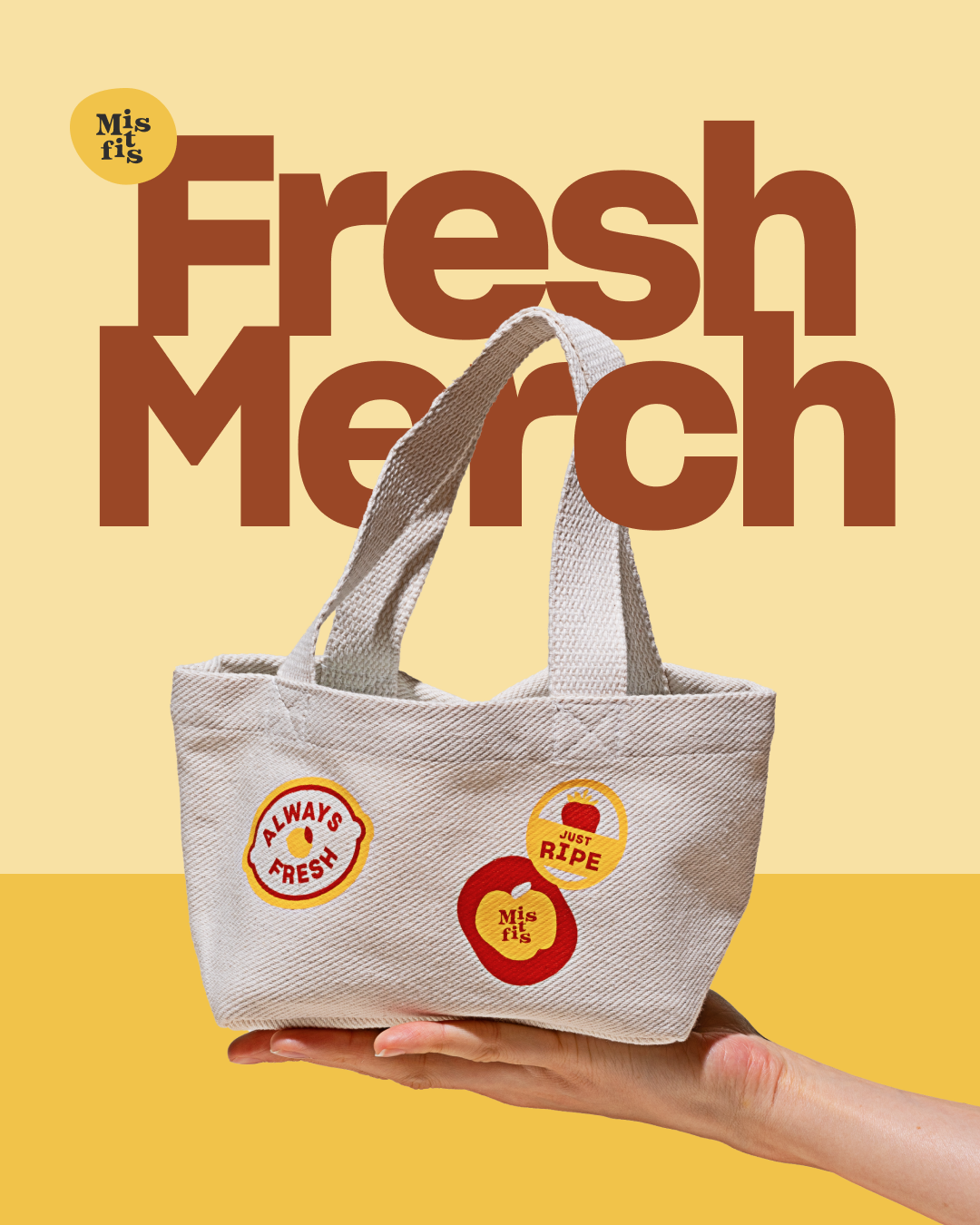 A hand holding a beige canvas tote bag with pepper and apple stickers. The background features large brown text reading "Fresh Merch" and smaller text "Misfits"