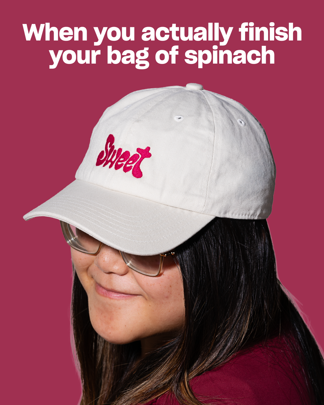 A person wearing glasses and a maroon shirt, with long dark hair, is looking down and smiling. They are wearing a white baseball cap with the word 'Sweet' embroidered in pink, against a maroon background, with white text above that reads, 'When you actually finish your bag of spinach.'