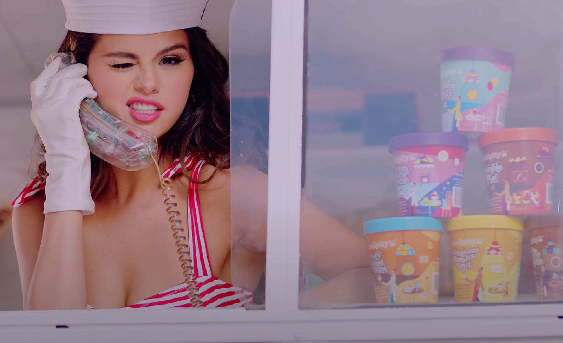 Selena Gomez in striped red and white top talking on a vintage telephone inside an ice cream truck, seen through a glass window displaying colorful containers of Serendipity ice cream.