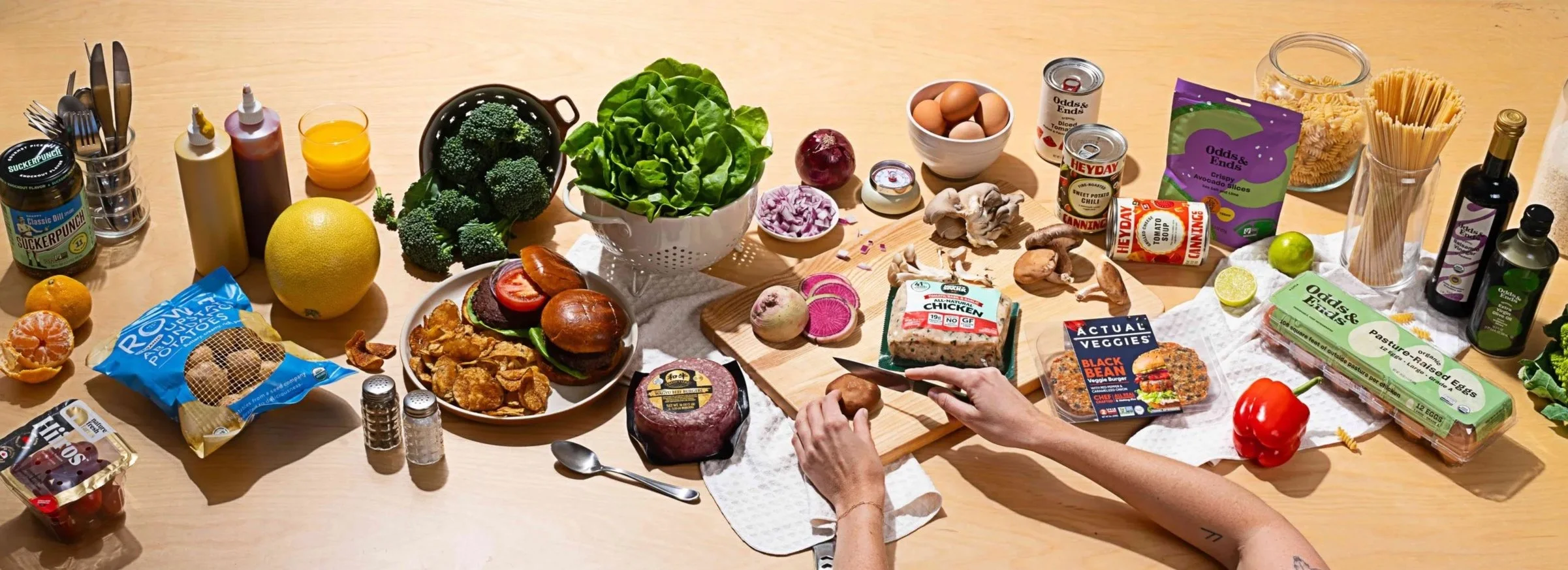 A wooden table filled with various groceries including vegetables, fruits, eggs, meats, packaged foods, condiments, and utensils, with a person preparing to cook.