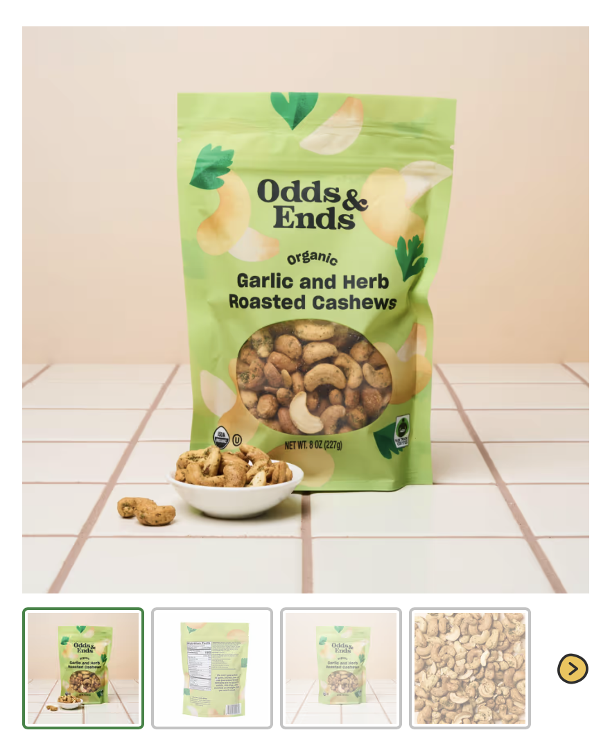 A bag of Odds & Ends organic garlic and herb roasted cashews standing on a tiled surface with a small bowl of cashews in front of it.