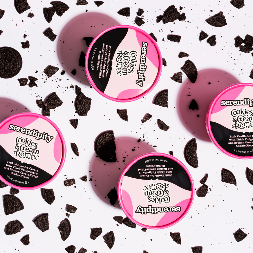 Three pink containers of Serenity Cookies & Cream Remix ice cream on a white surface with crushed Oreo cookies scattered around.