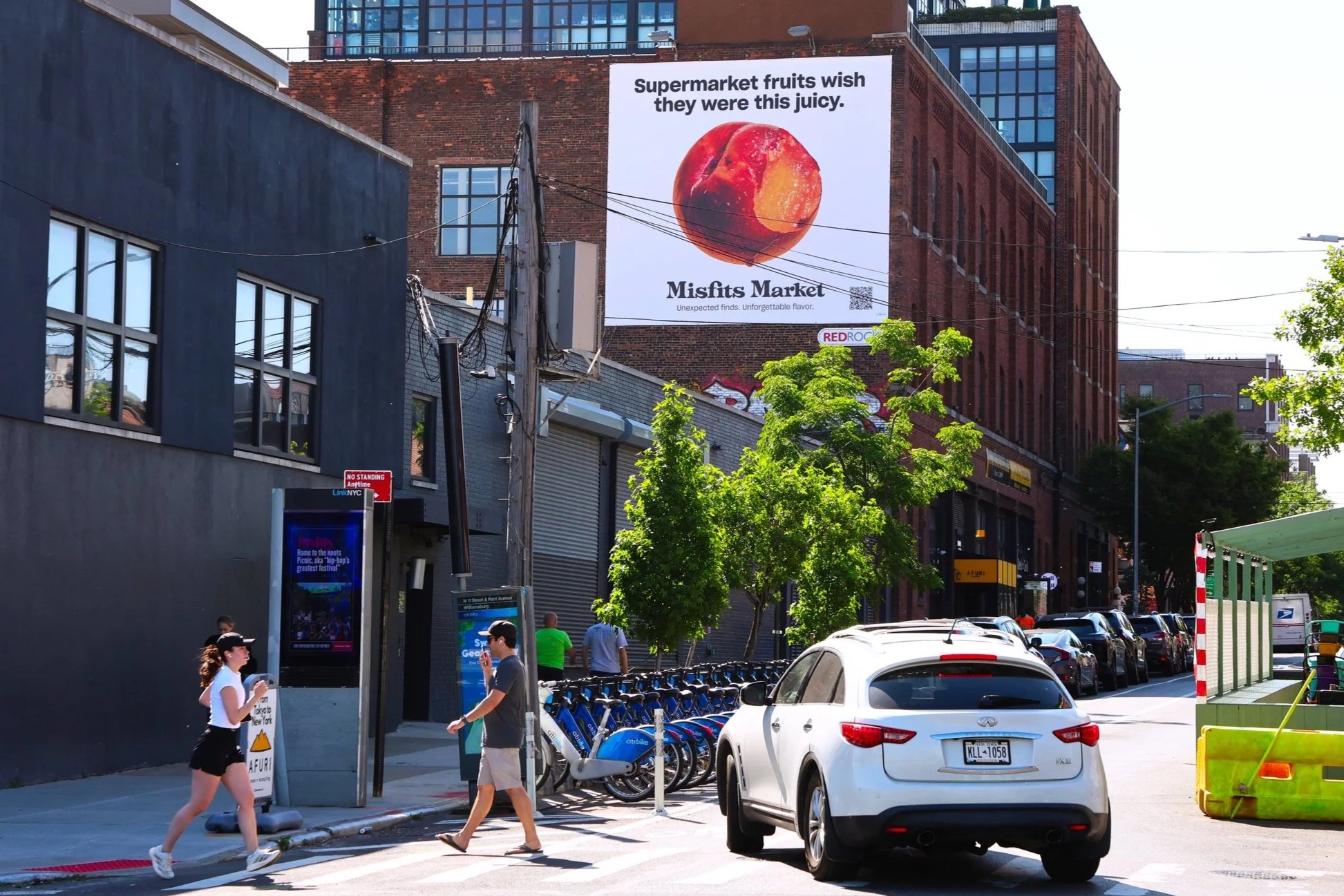 Street scene with pedestrians, parked cars, and a large billboard showing a peach with the text 'Supermarket fruits wish they were this juicy. Misfits Market.' Trees line the sidewalk, and a bike-sharing station is visible.