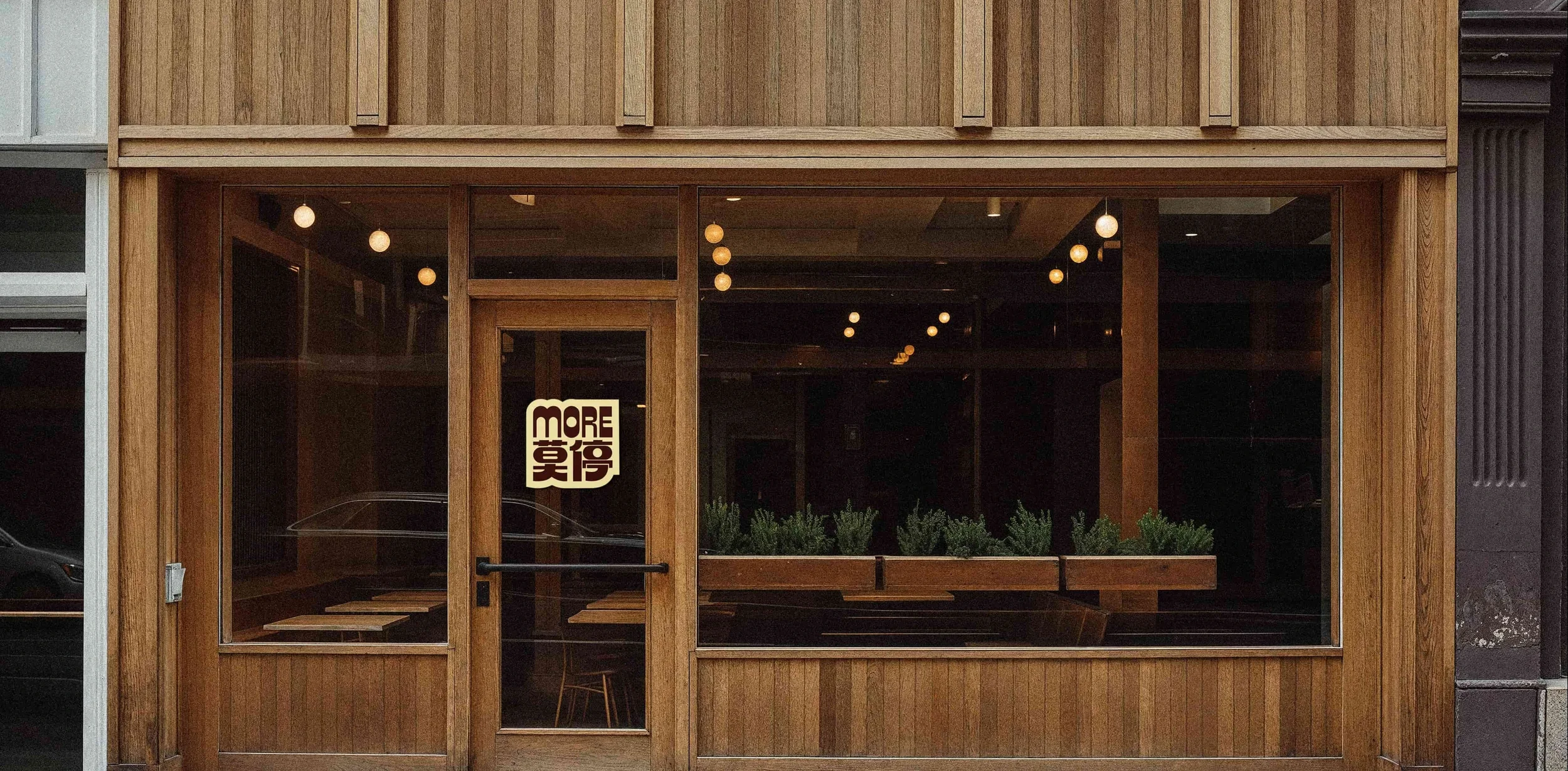 A wooden storefront with large glass windows displaying a sign that says "MORE" and some Chinese characters, with interior lighting and greenery visible inside.