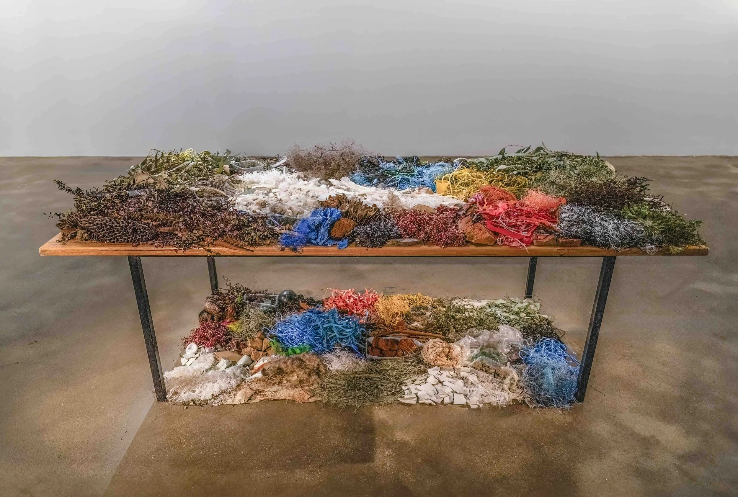 Installation of colourful found materials, grouped by colour and arranged on the table top and below in the rectangle beneath the table.