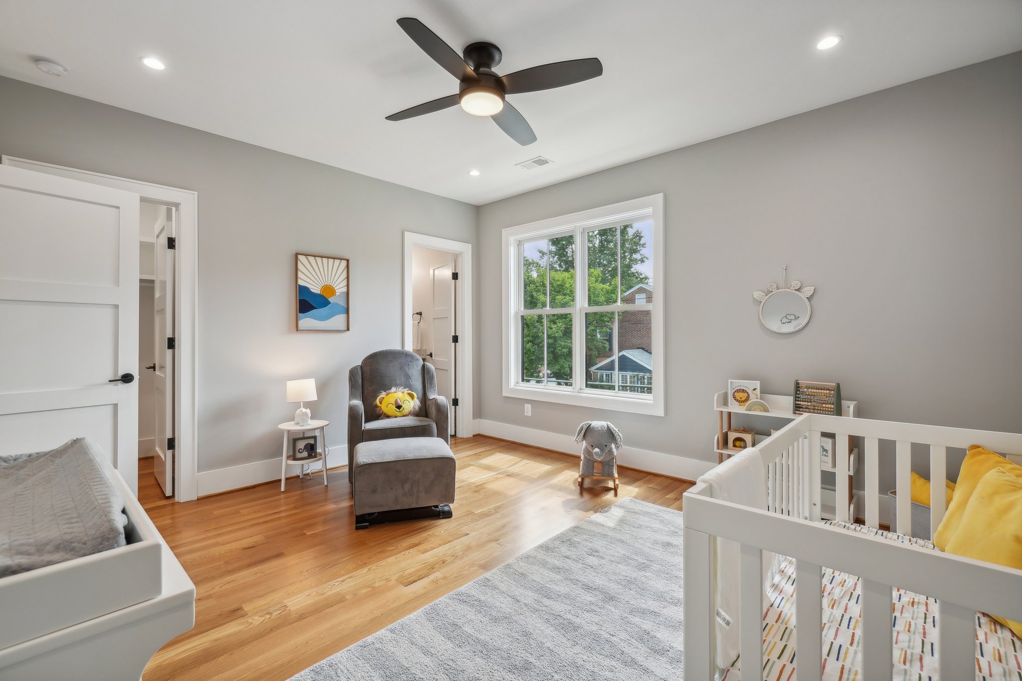 Modern nursery with large window, and ceiling fan. Features include a white crib with yellow bedding, framed animal art, a gray armchair with ottoman, wooden floor, and a rocking horse.