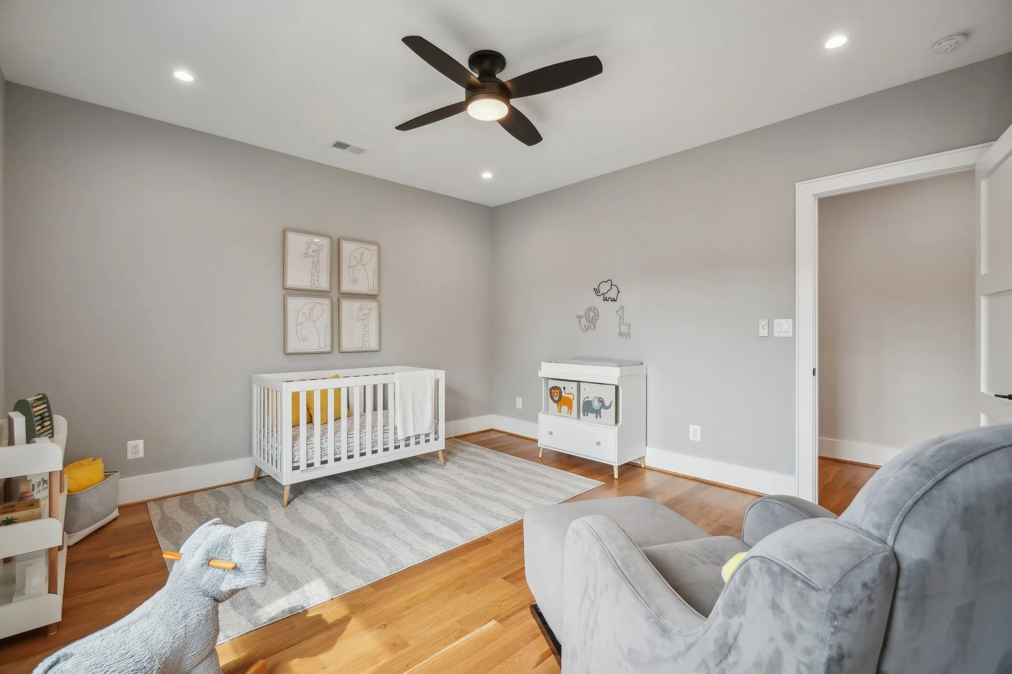 Modern nursery with large window, and ceiling fan. Features include a white crib with yellow bedding, framed animal art, a gray armchair with ottoman, wooden floor, and a rocking horse.