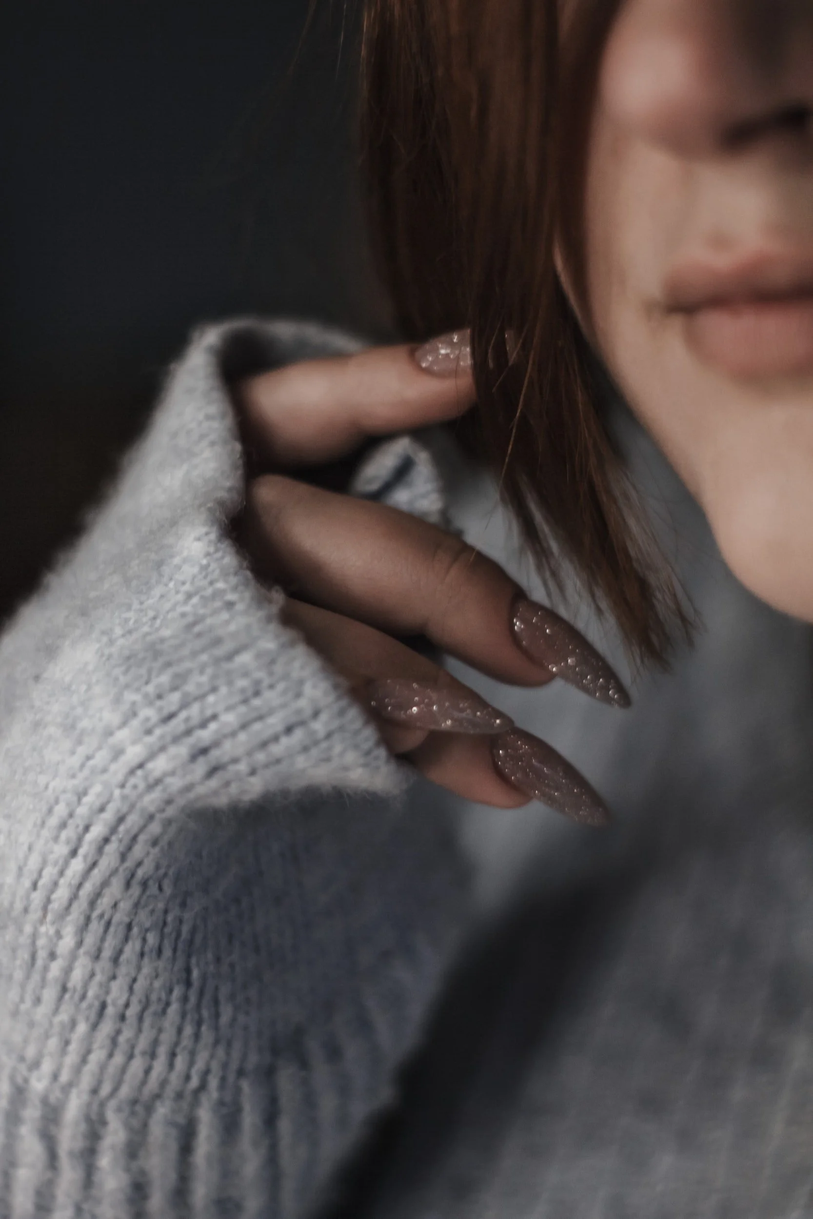 Close-up of a woman with brown hair, wearing a grey sweater, touching her hair near her ear, with glittery long nails visible.