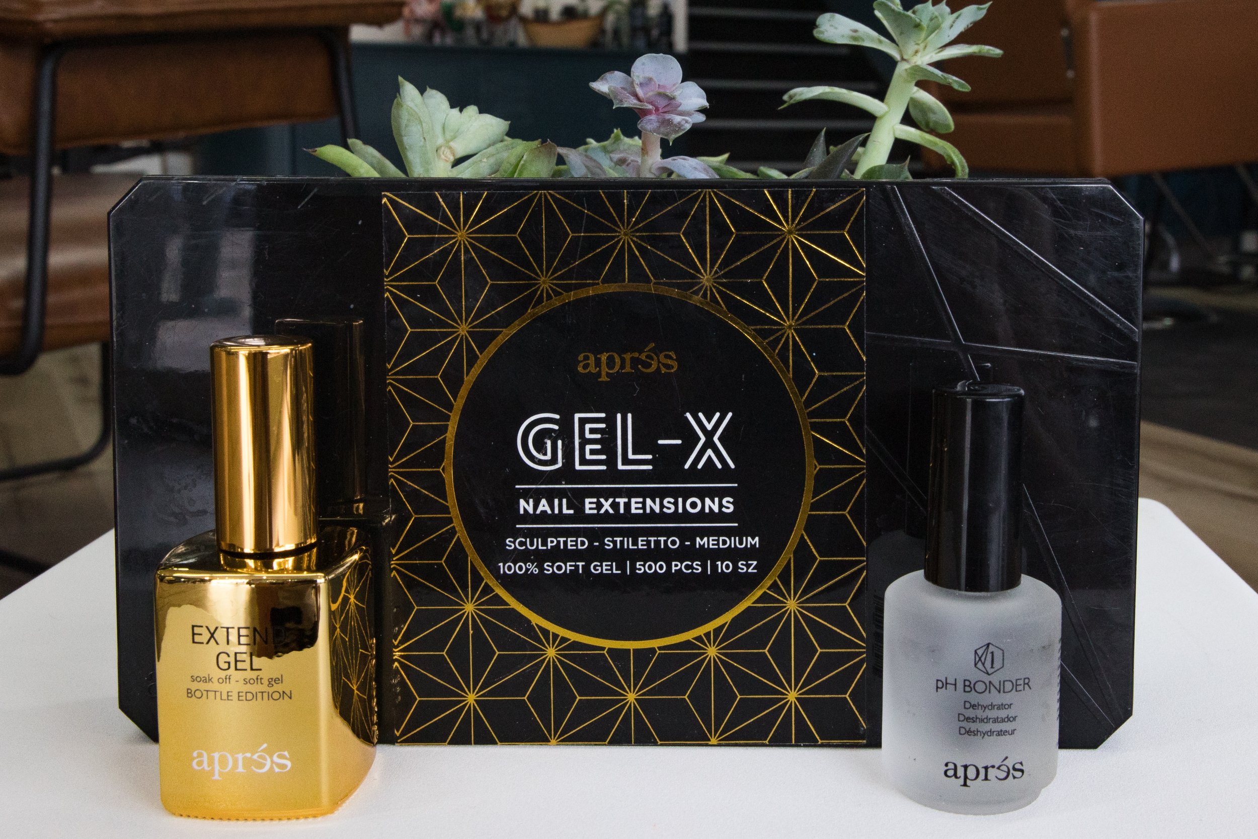 A set of nail extension gel products including a bottle of soak-off gel, a black box labeled 'GEL-X', and a white bottle labeled 'PH Bond', with a potted succulent plant in the background.