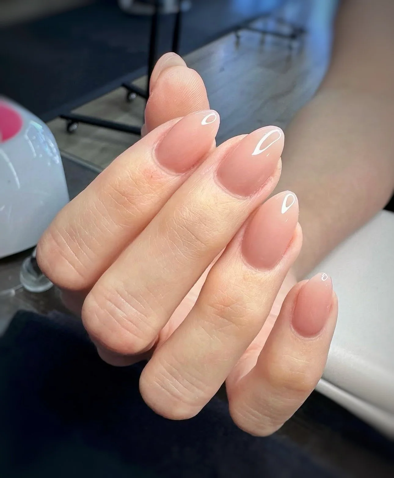 Close-up of well-manicured nails with clear polish and white accents, held in front of a blurred background with a palette and table.