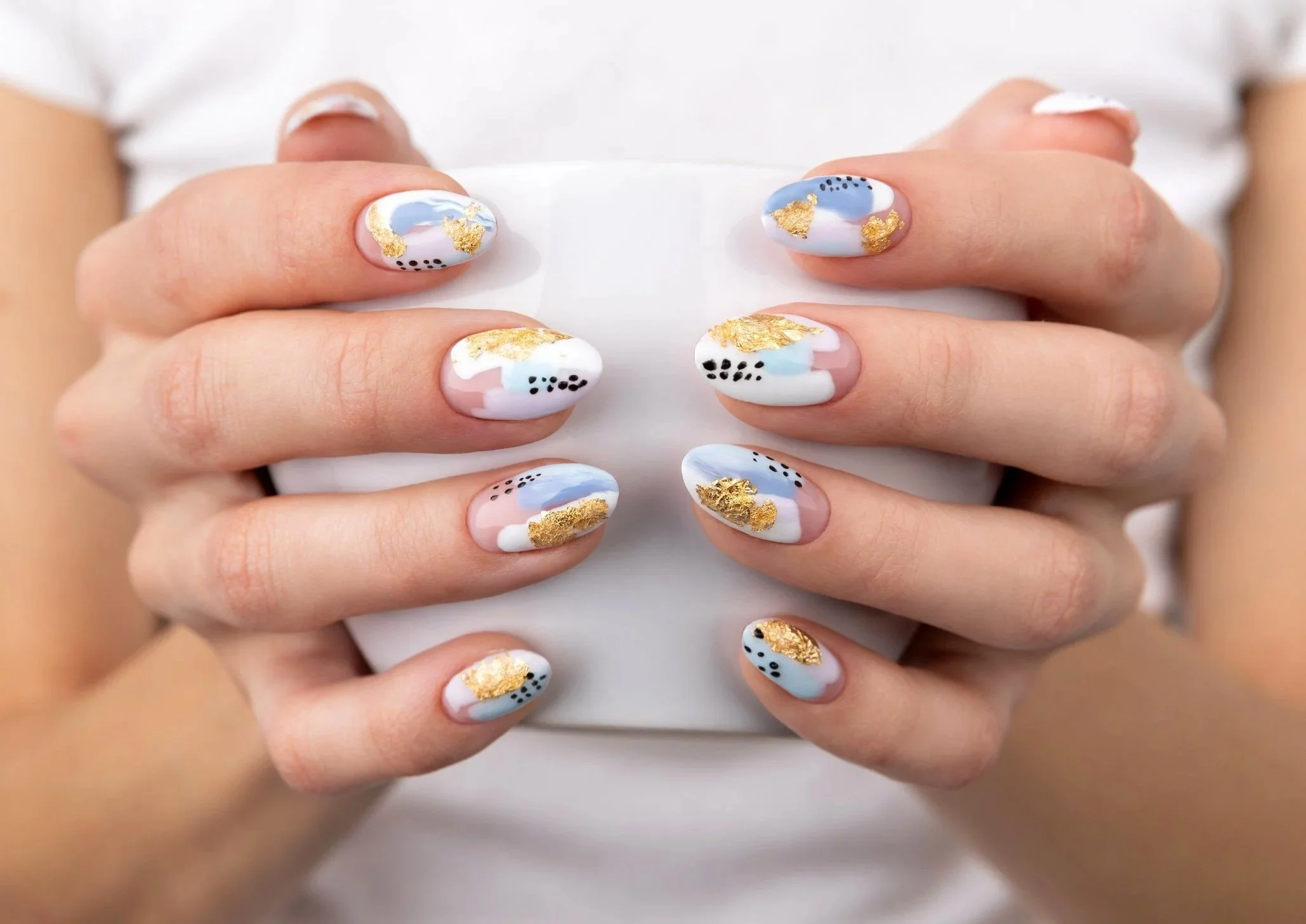 Close-up of hands with decorated nails featuring abstract pastel blue, white, pink, black dotted, and gold foil designs.