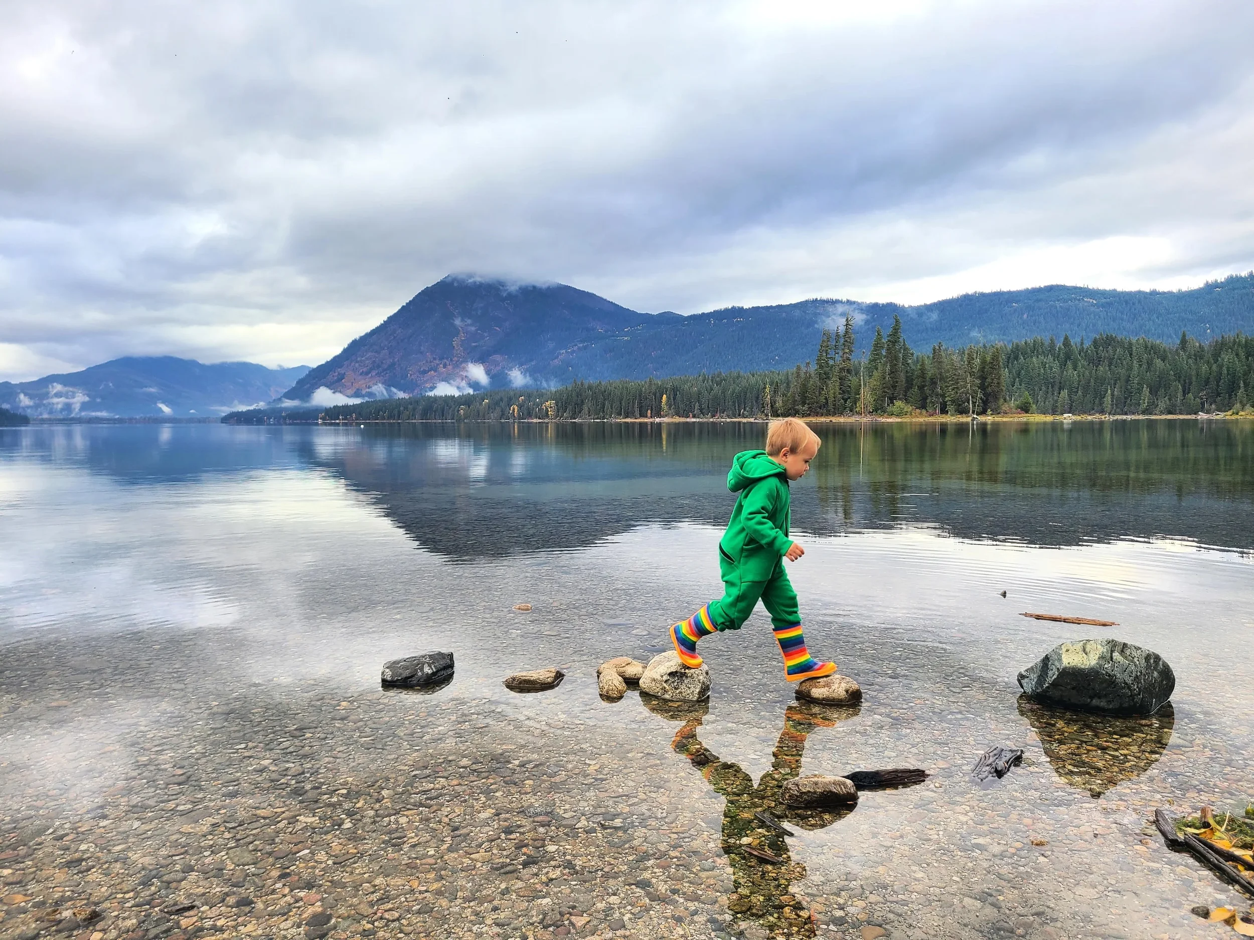 Child wearing a green zip-up Zippy Zips suit and rainbow boots, walking across rocks in a picturesque lake mountain scene