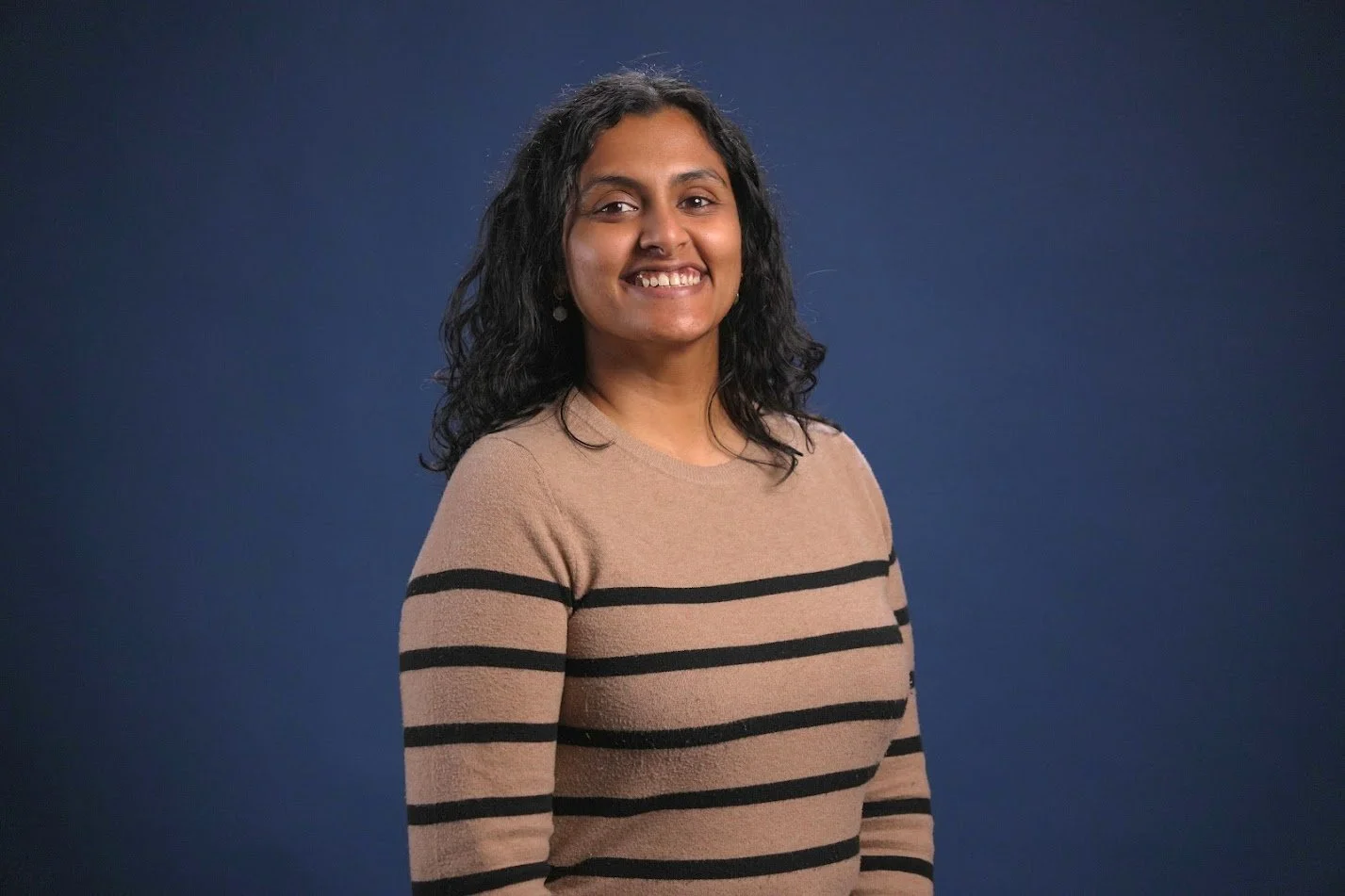 Headshot of Sandhya Simhan, a woman with long curly dark hair smiling wearing a tan sweater with black stripes