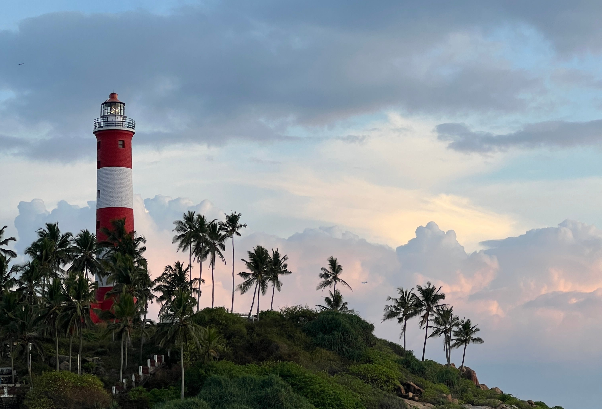 A tall red and white striped lighthouse stands amidst lush green palm trees under a cloudy sky during sunset or dusk.