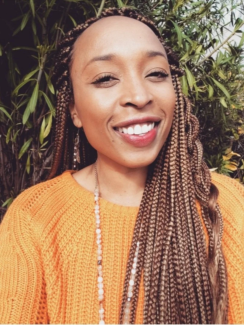 Person smiling in an orange sweater with braided hair, outdoors with greenery in the background.