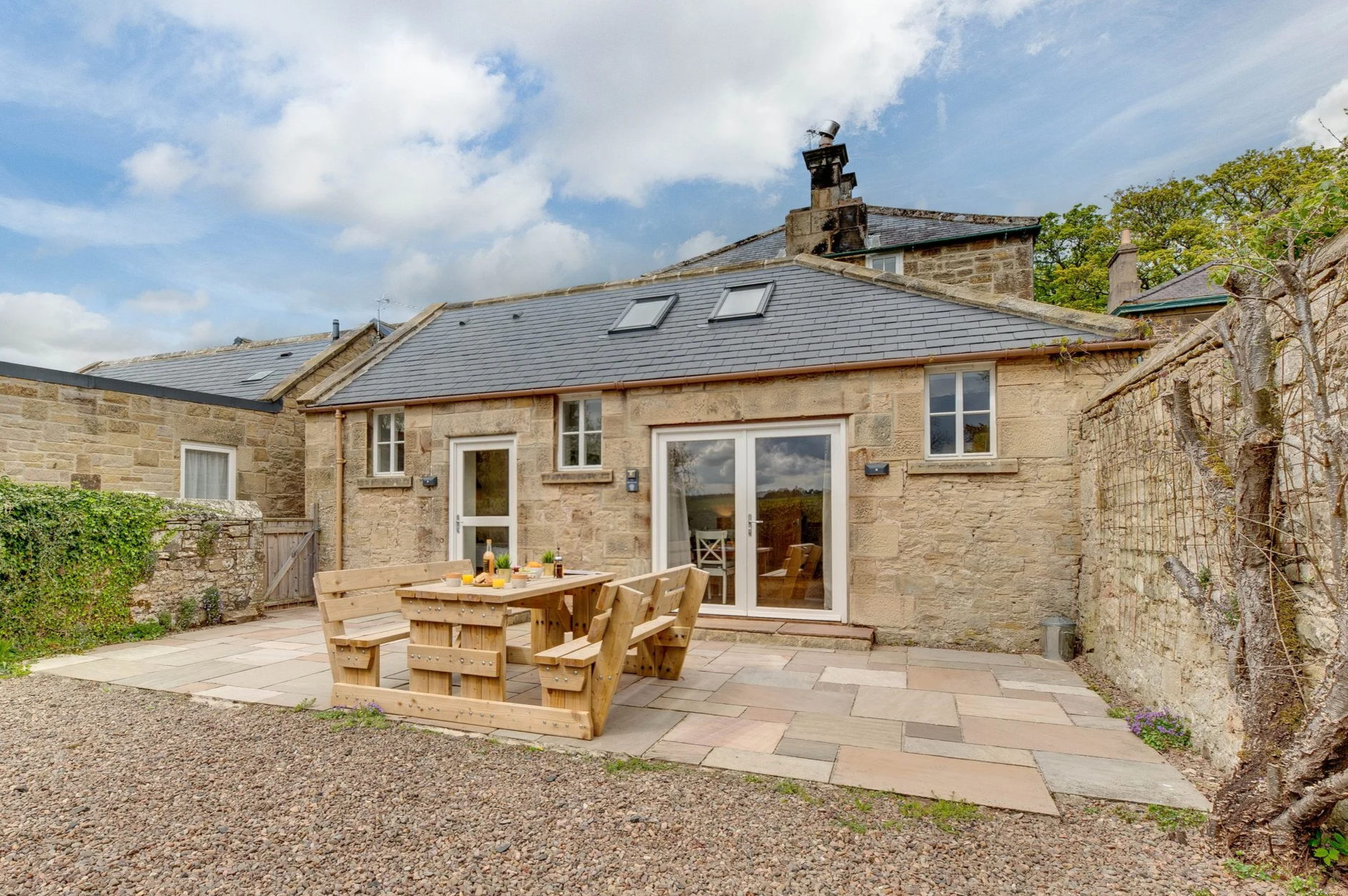 Stone house with slate roof, patio with wooden dining table and chairs, sliding glass door, small windows, and a stone wall with ivy on the left.