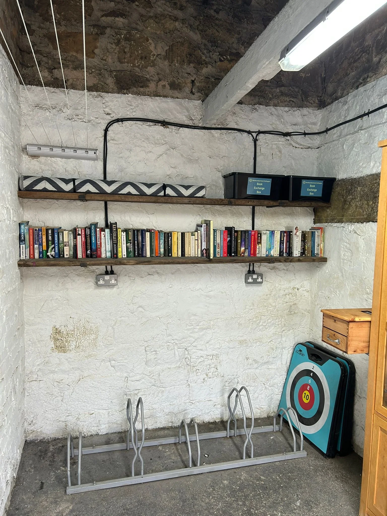 Indoor scene with three wooden shelves filled with books, electrical outlets and cords on the wall behind, a target practice board leaning against the wall, and metal bike racks in the foreground.