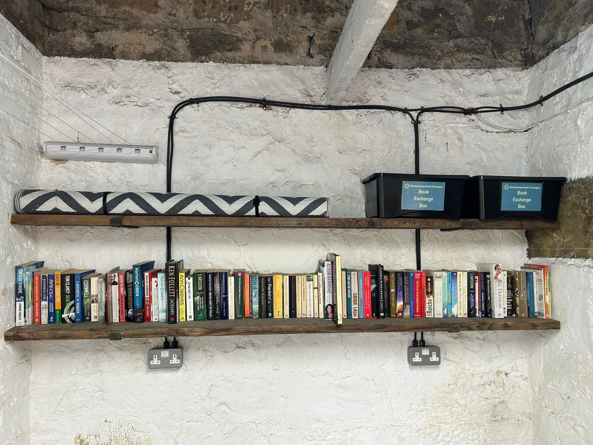 Two bookshelves with a collection of books, wall with white textured paint, and electrical outlets with plugs. The upper shelf has a rolled-up black and white patterned mat and a black box labeled 'Book Exchange Box'.