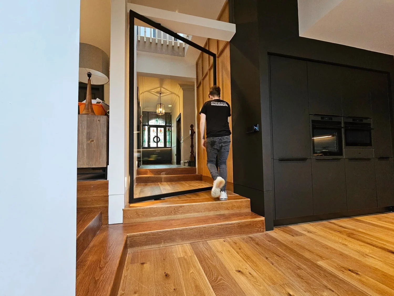 A person walking down a hallway with herringbone flooring past a tall black steel framed pivot door beside a dark fitted kitchen.