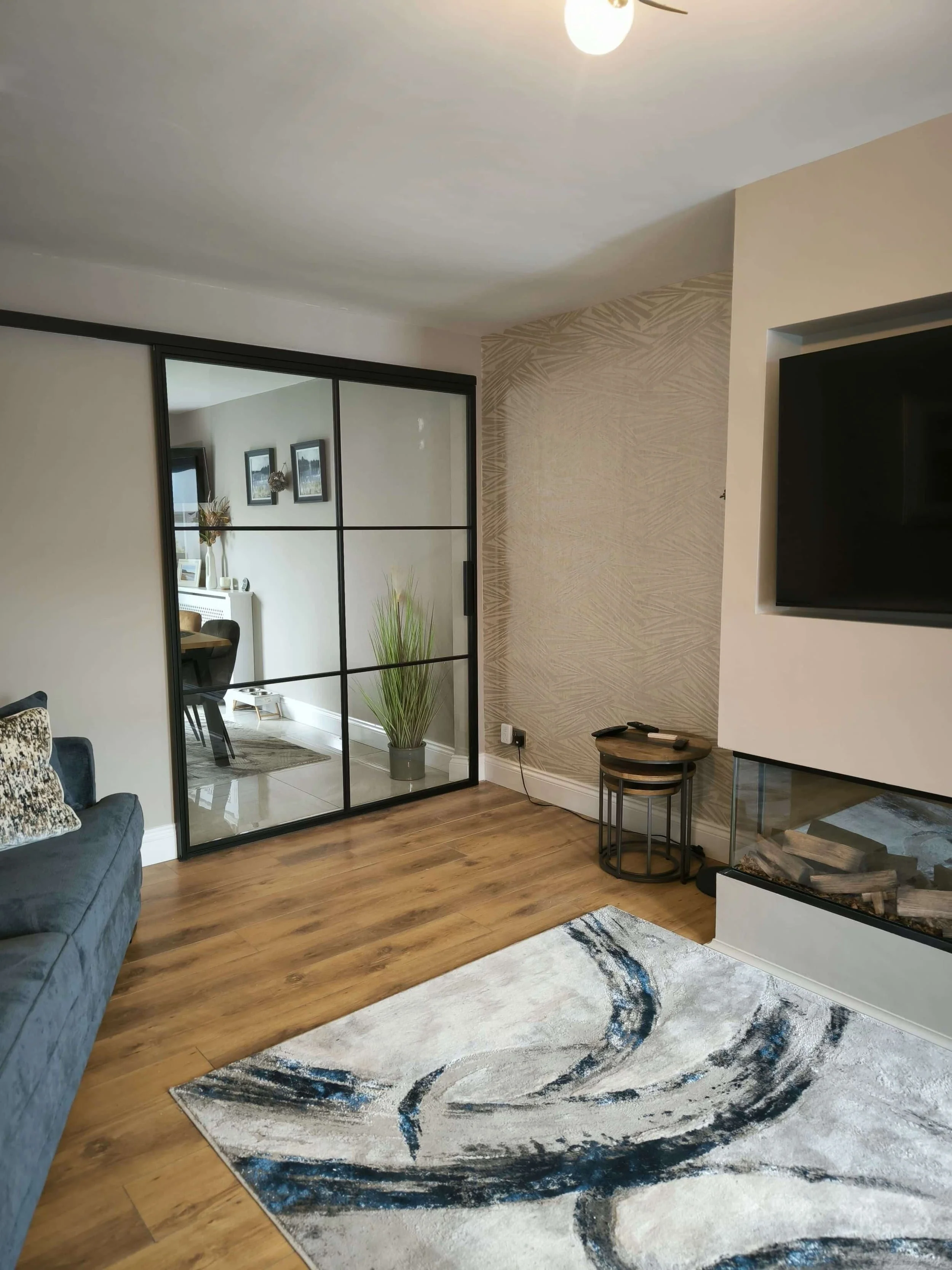 Modern living room with a blue sofa, area rug, wall-mounted TV, fireplace, and glass sliding doors reflecting a dining area.