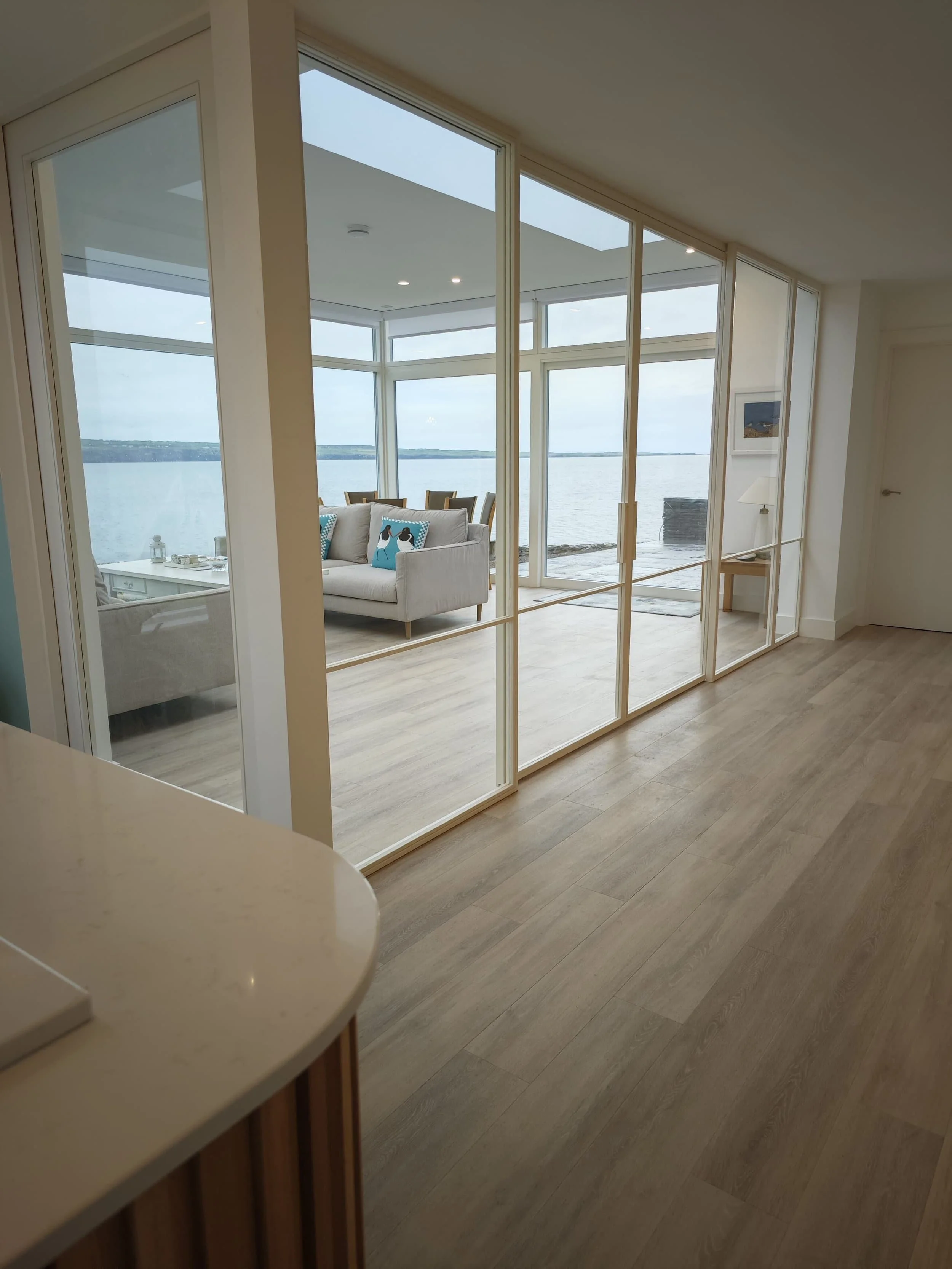 Modern living room with glass walls, ocean view, light wood floors, and a cozy seating area with blue accent pillows.