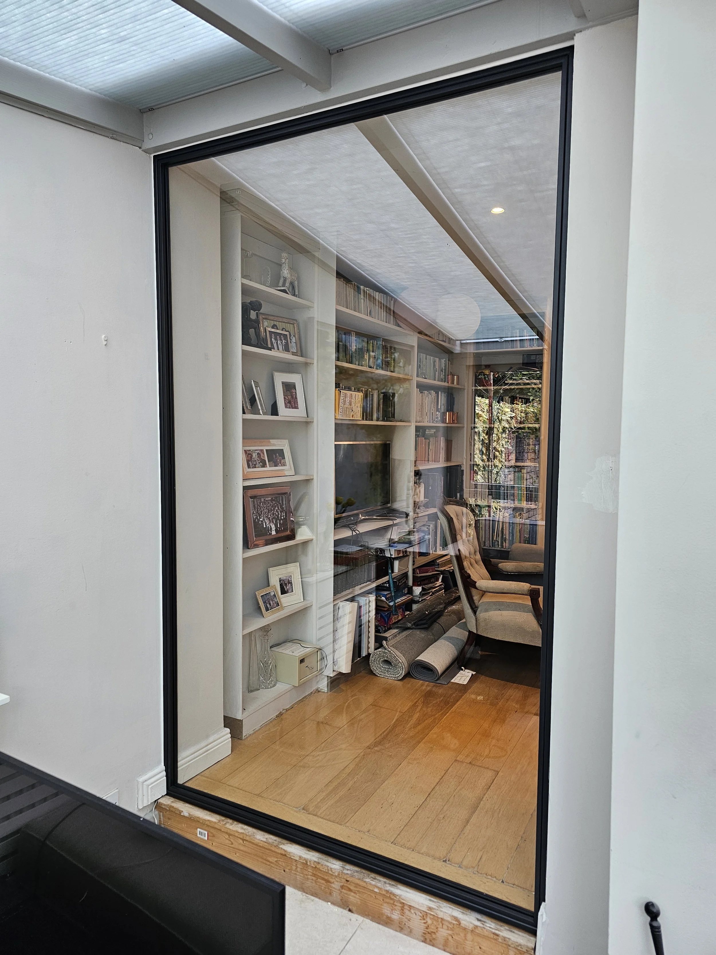 View through a glass door into a living room with bookshelves, a television, chairs, and rolled-up rugs on a wooden floor.