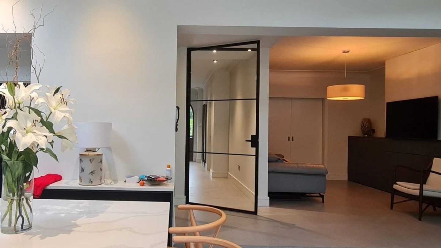 A white hallway with a black steel framed hinged glass door standing open into a warm lamplit living room, lilies on the counter in the foreground.