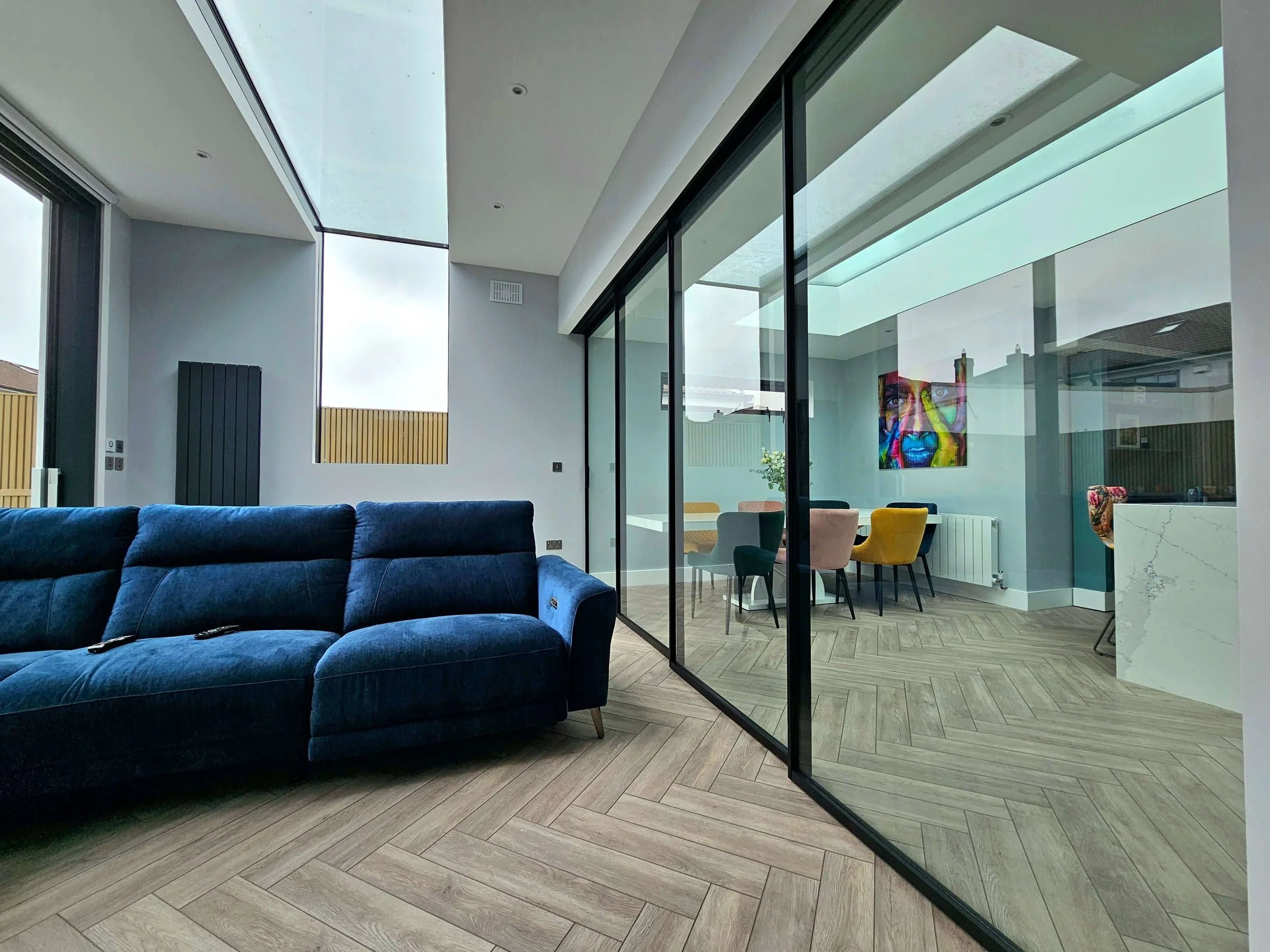 Modern living room with a blue sofa, glass wall, and view into a bright dining area with colorful chairs and abstract art.