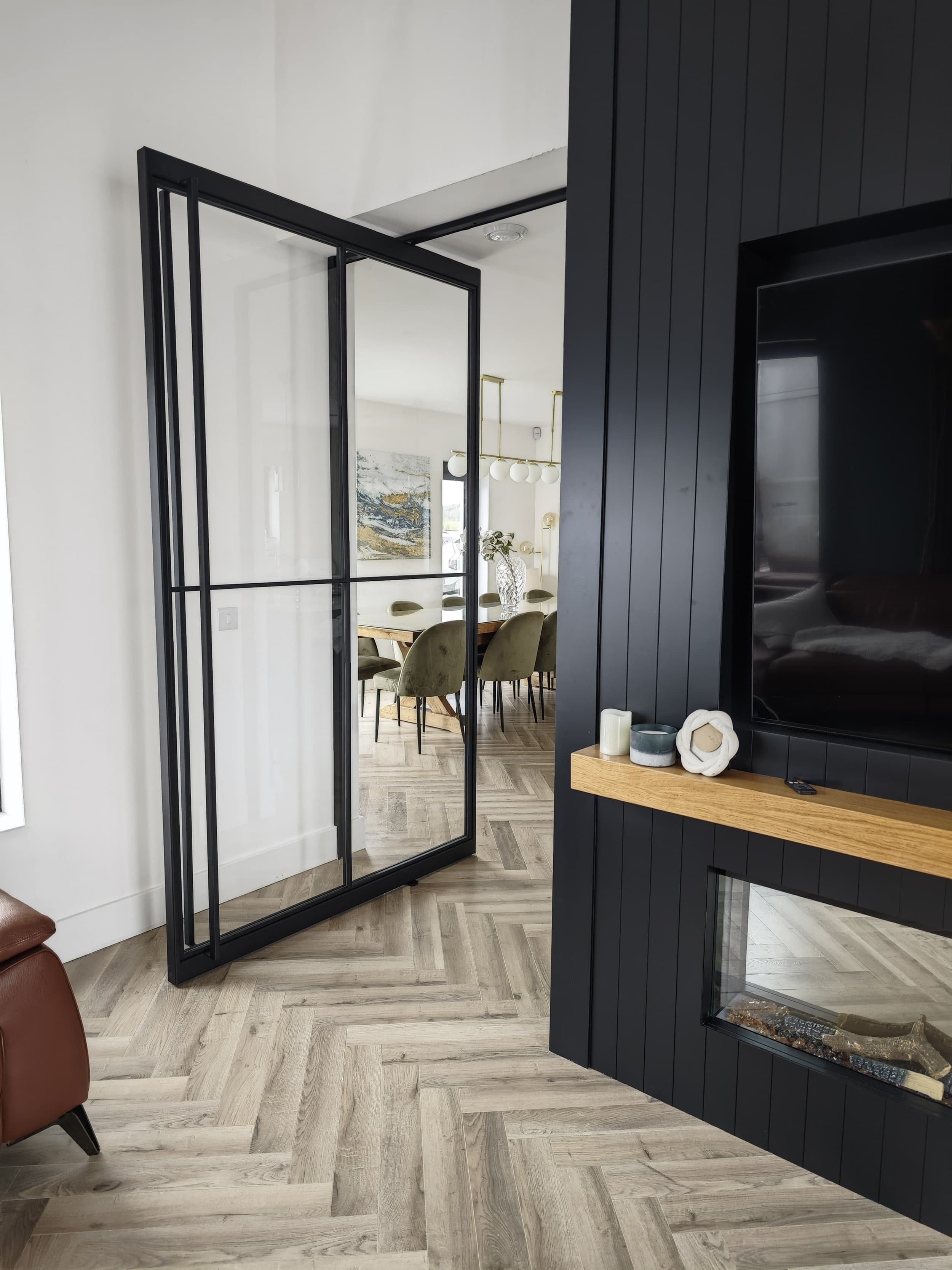 Modern glass door opens to a dining area with green chairs; black wall, wood shelf, and fireplace in the foreground.