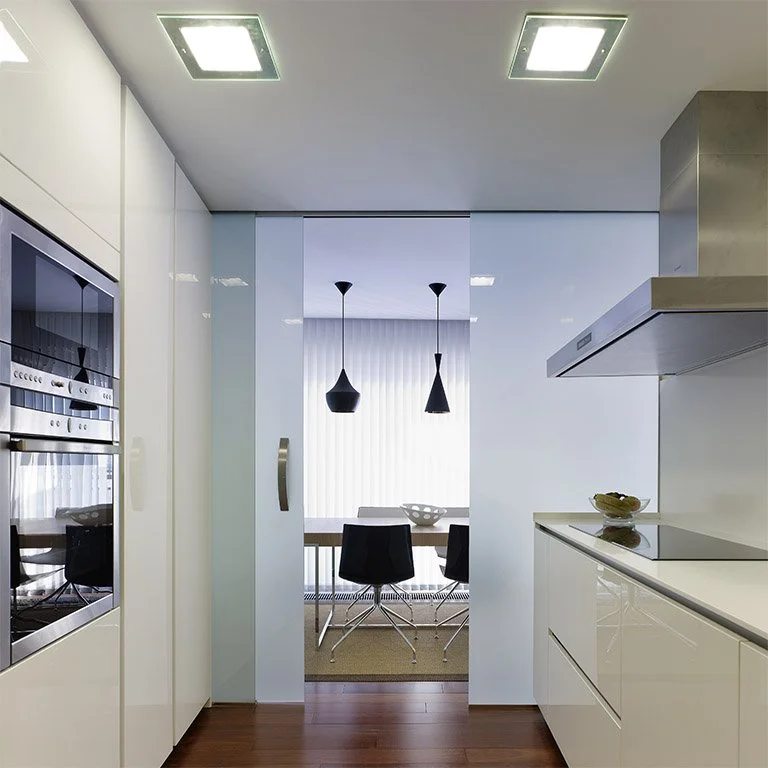 Modern kitchen with built-in oven and microwave, white cabinets, a stove, a vent hood, and a dining area with a table and black chairs seen through a sliding glass door.