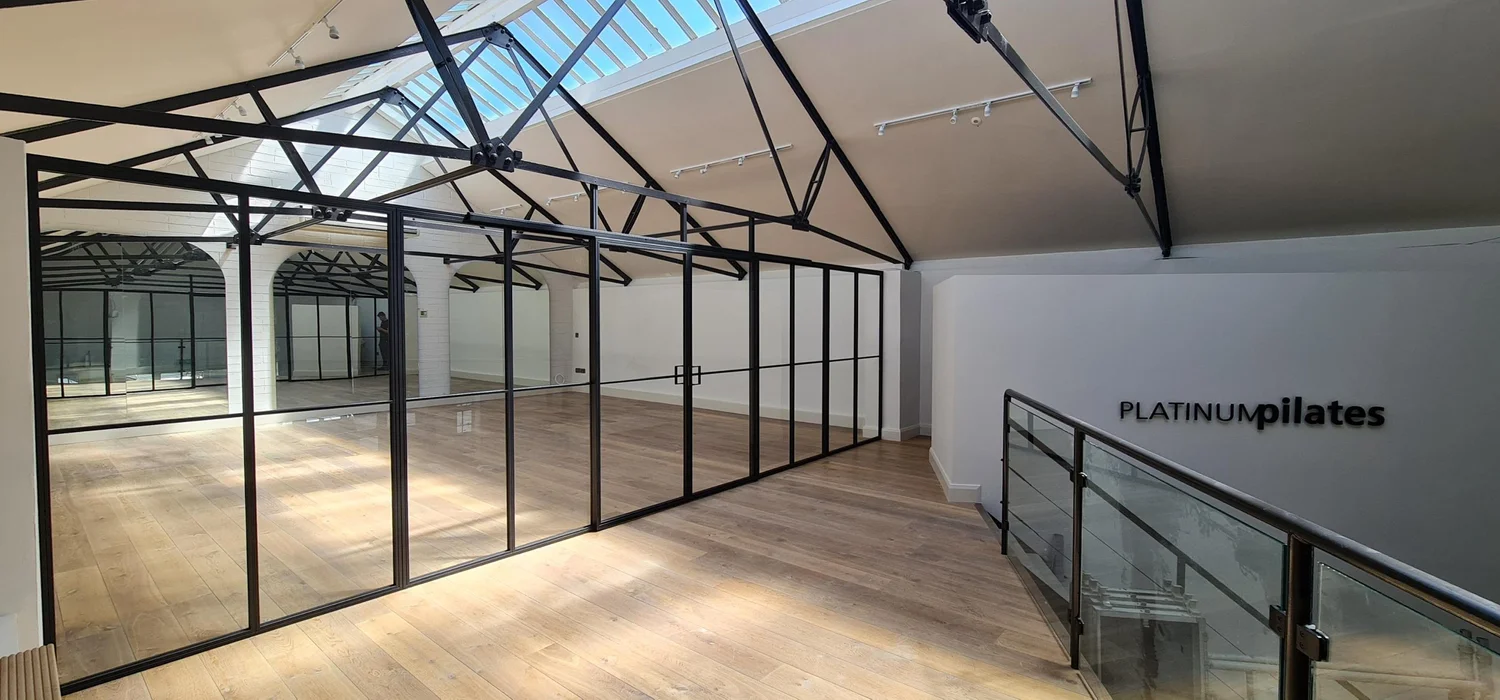 A first floor studio under a pitched rooflight, divided along its length by a black steel framed glass partition with sliding doors, with Platinum Pilates lettering on the far wall.