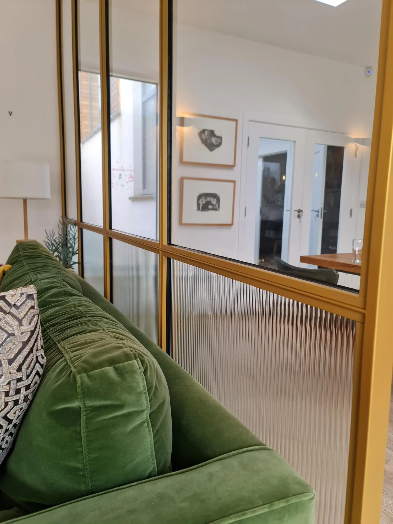 A living room with a green velvet sofa in front of a brass framed steel and glass partition with fluted glass, looking through to a hallway.