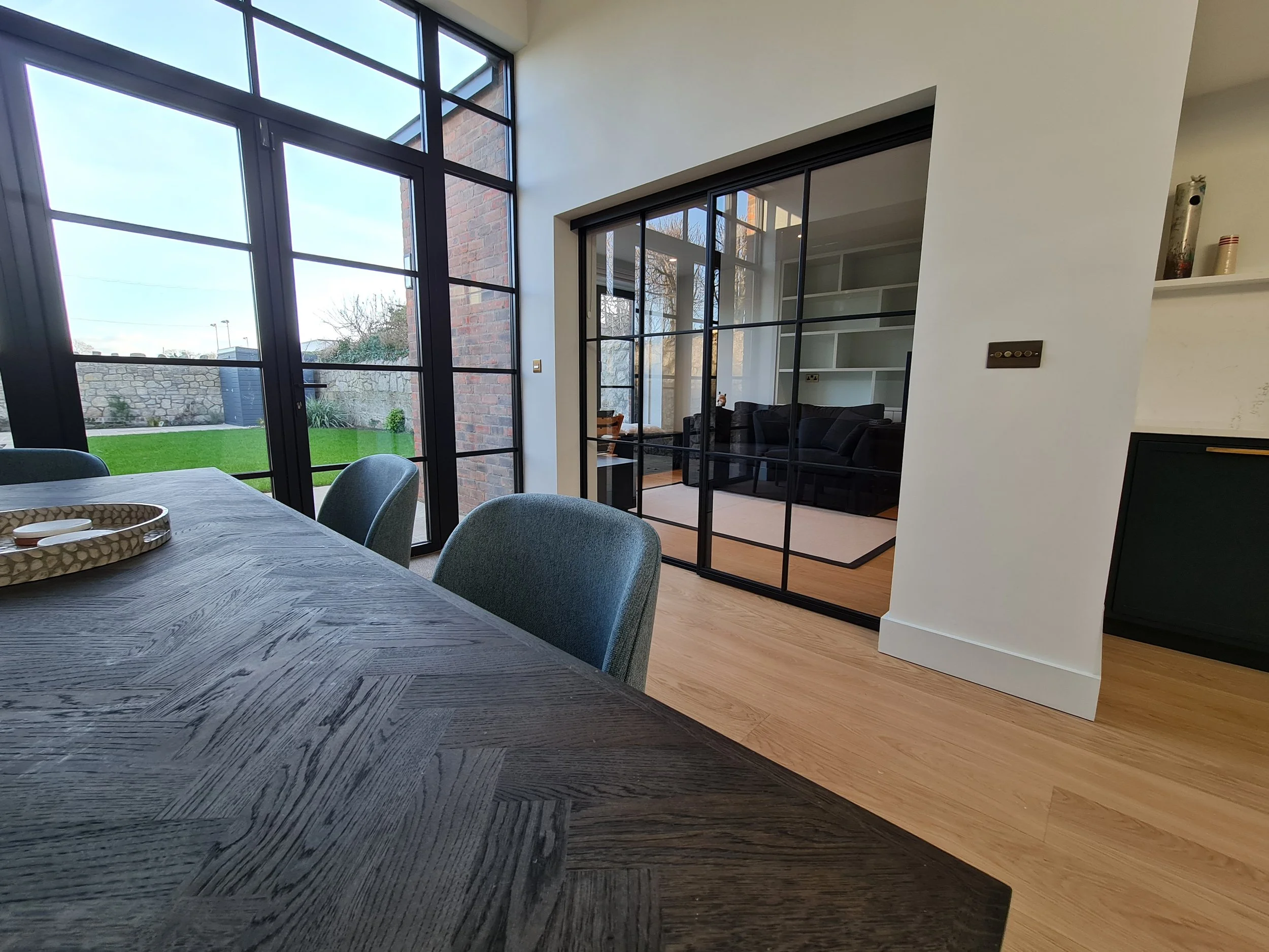 A dining room with a black steel framed glazed wall onto the garden, and a black steel framed sliding partition on the right looking through into a living room.