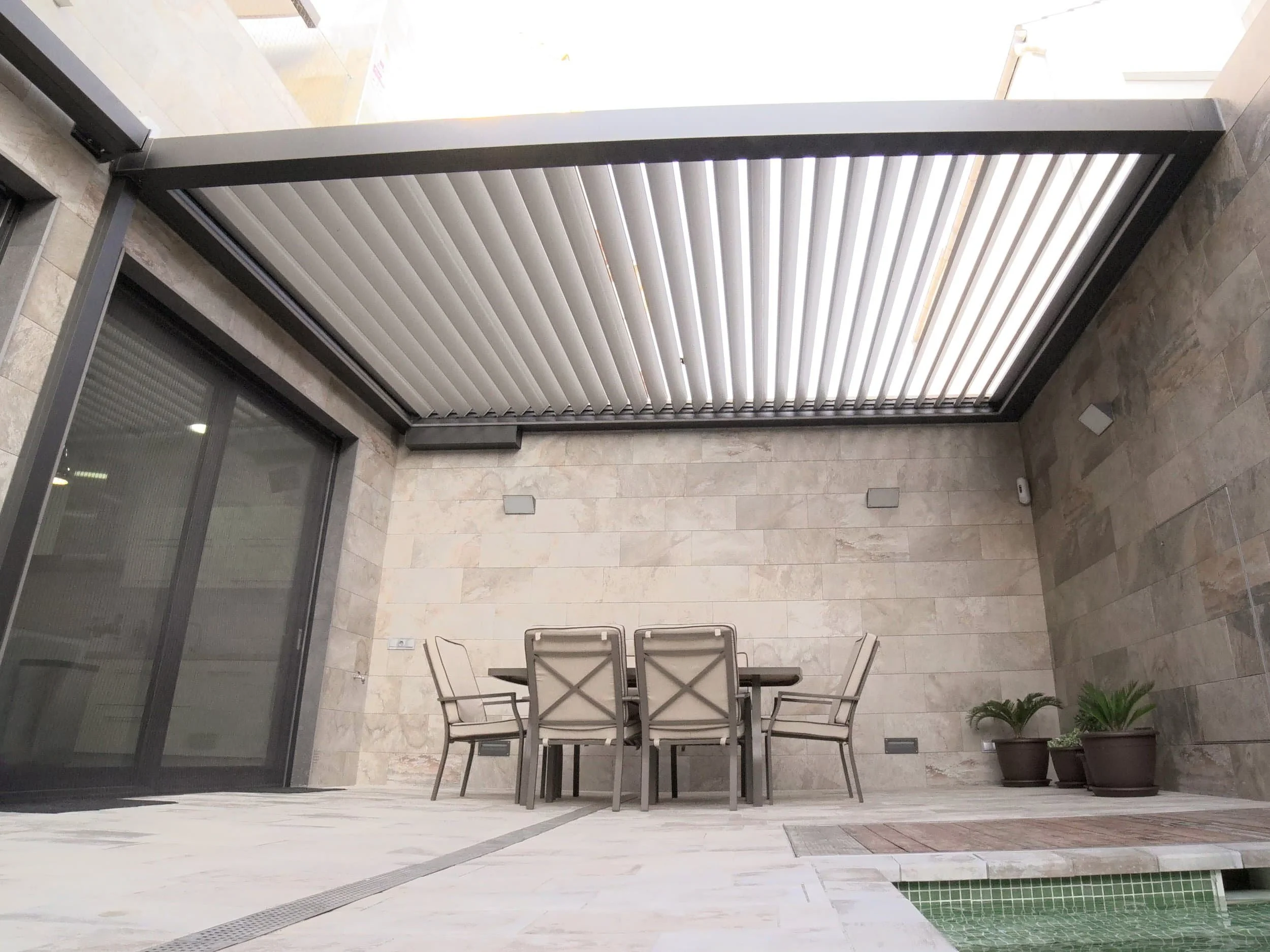 Outdoor patio with a slatted pergola roof, dining table and chairs, potted plants, and stone tile walls and flooring.