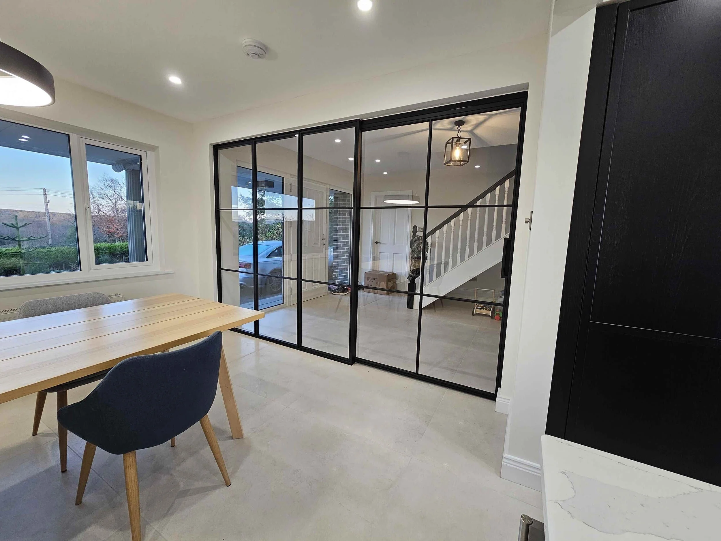 Modern dining area with wooden table, blue and gray chairs, and glass sliding doors revealing a hallway and staircase.