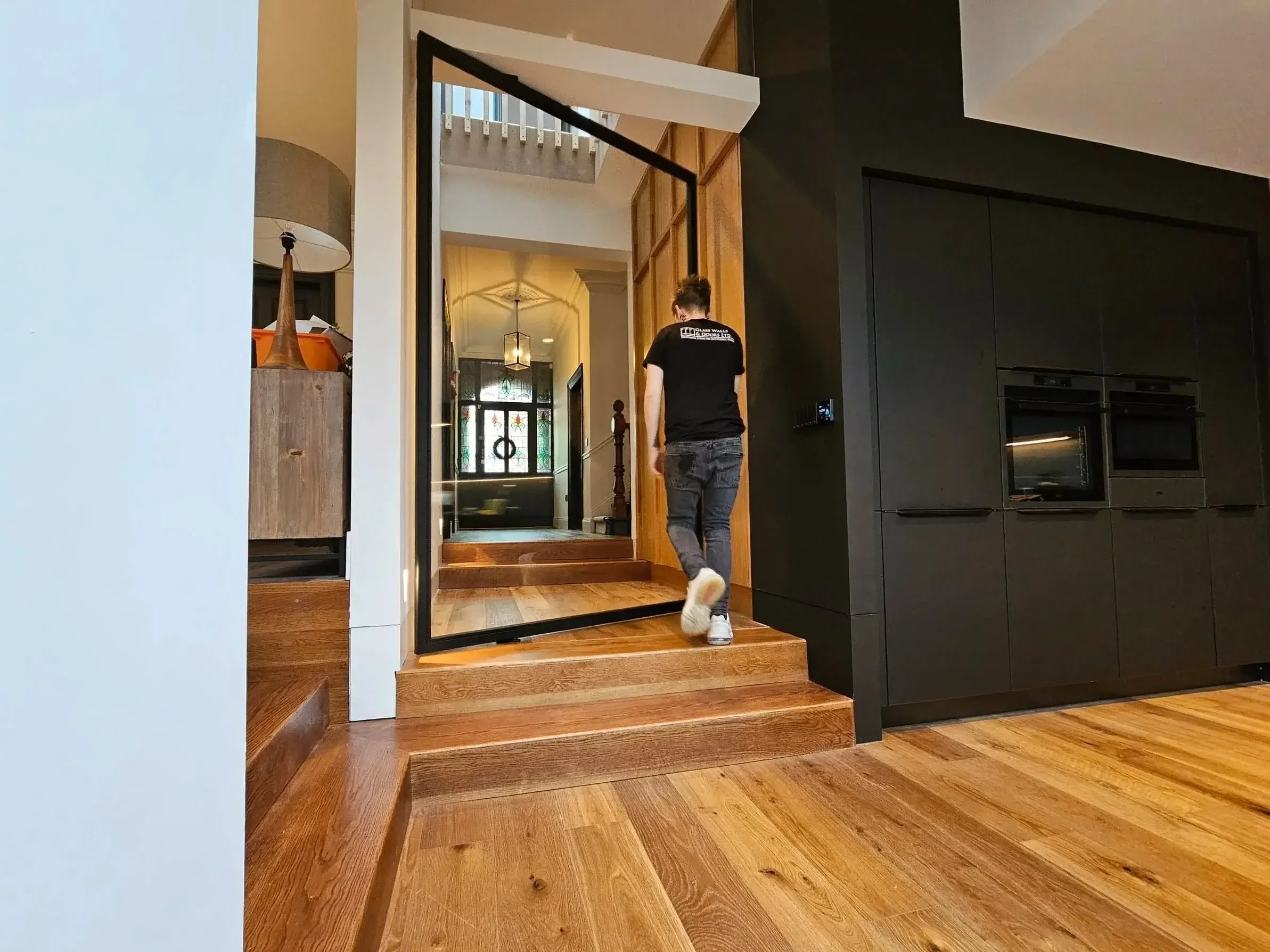 A person walking on wooden stairs inside a modern home, with a black wall featuring an integrated oven, and a cozy entrance area with stained glass door in the background.