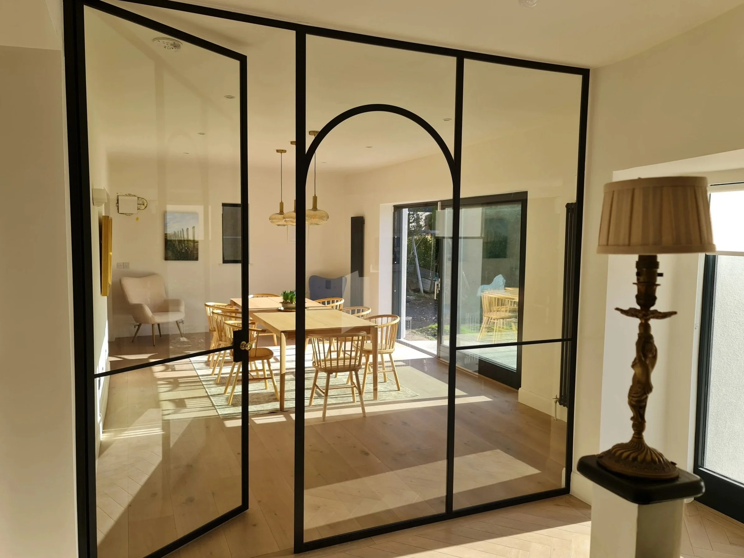 Glass partition with black frame and arch separates a bright dining area with a wooden table, chairs, and pendant lights.
