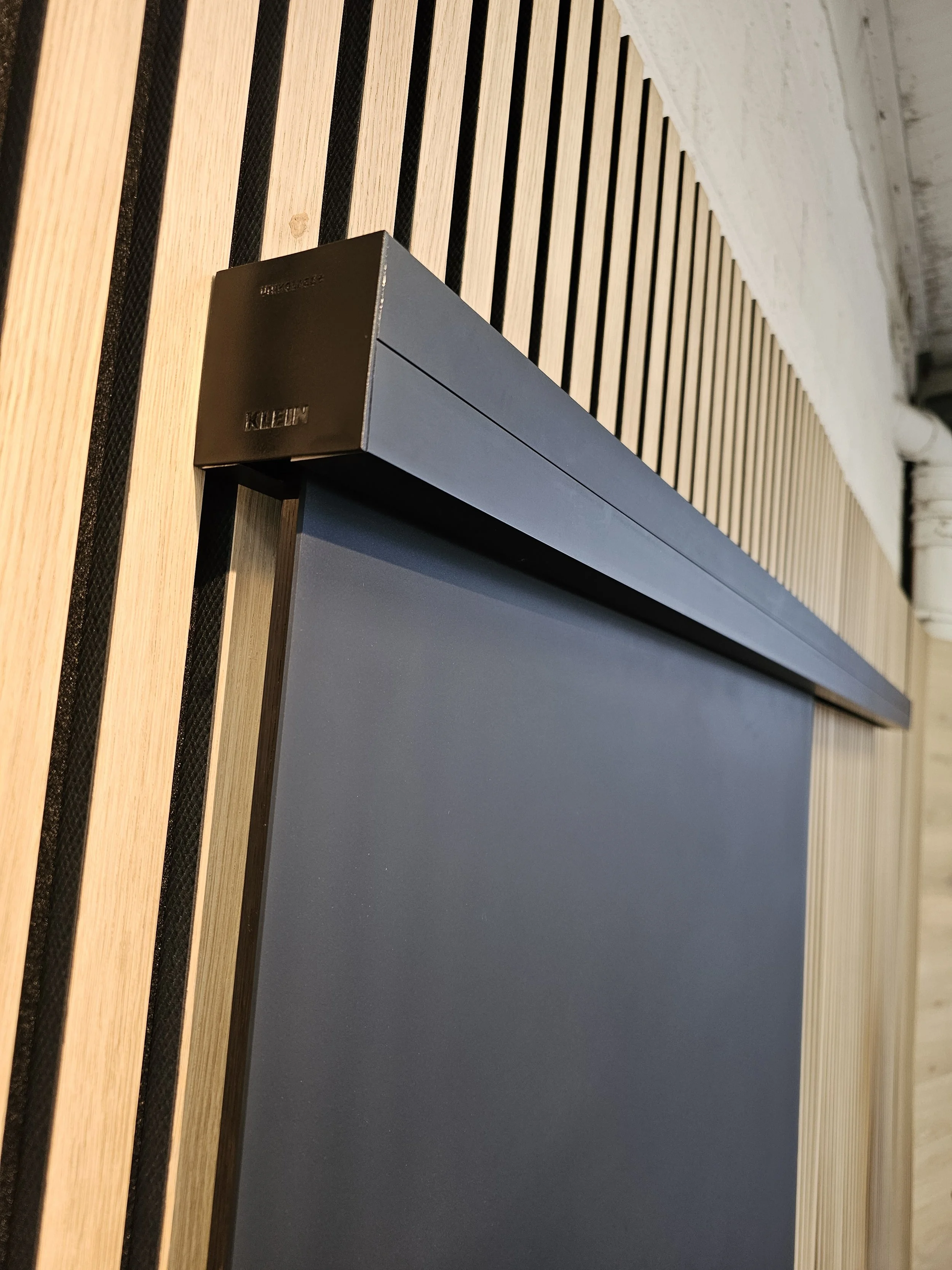 Close-up of a black Klein door frame with the top corner showing, against a background of vertical light wooden slats and a white brick wall.