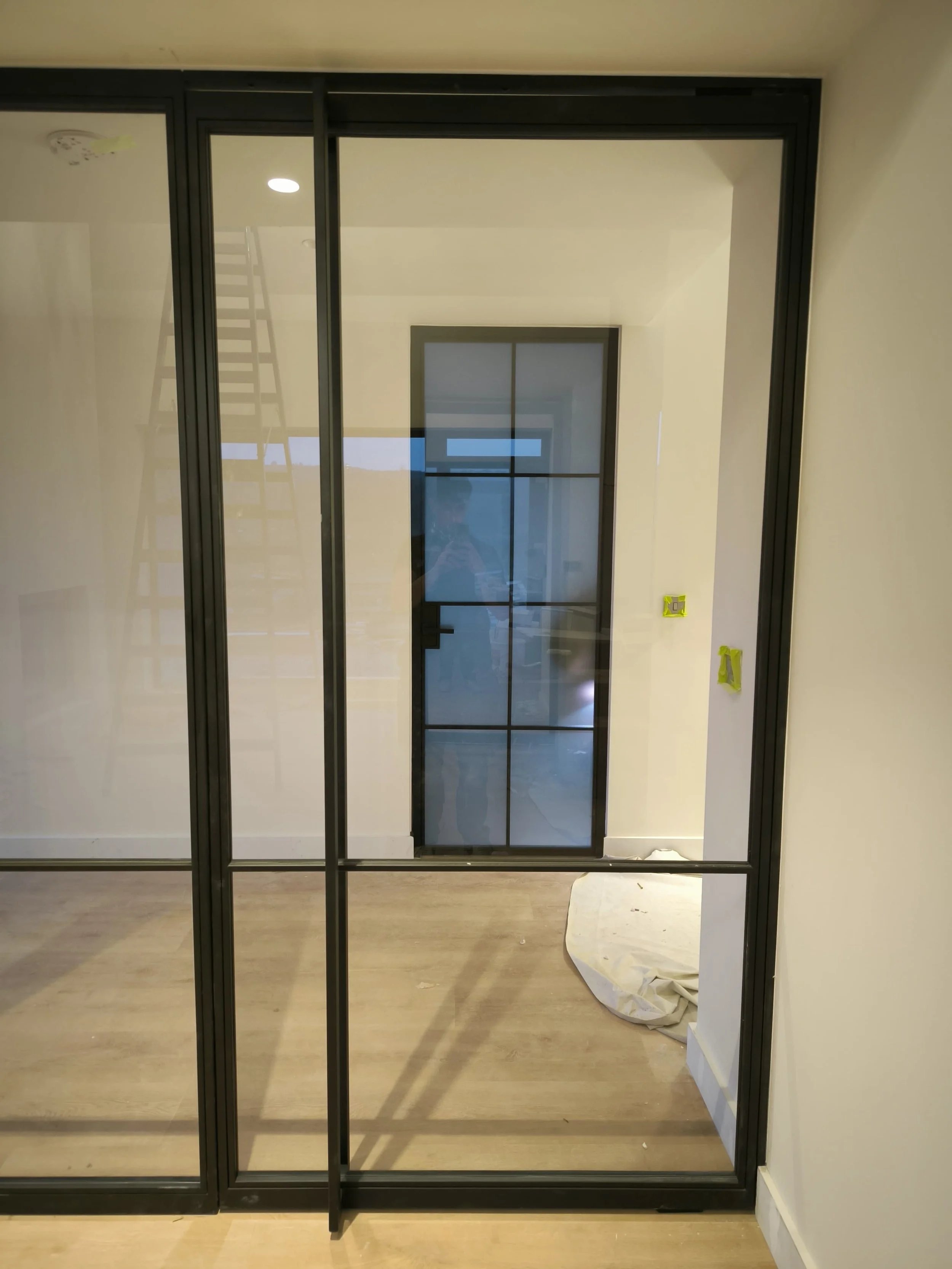 Glass partition with black frame in a modern room, a ladder and door with black grid visible in the background.